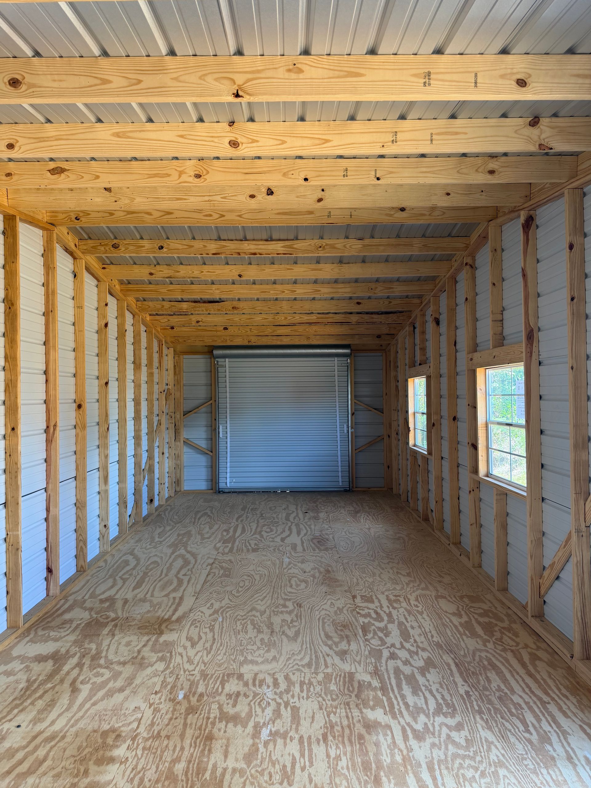 Empty unfinished shed interior with exposed wood framing, plywood floor, and a roll-up door at the far end
