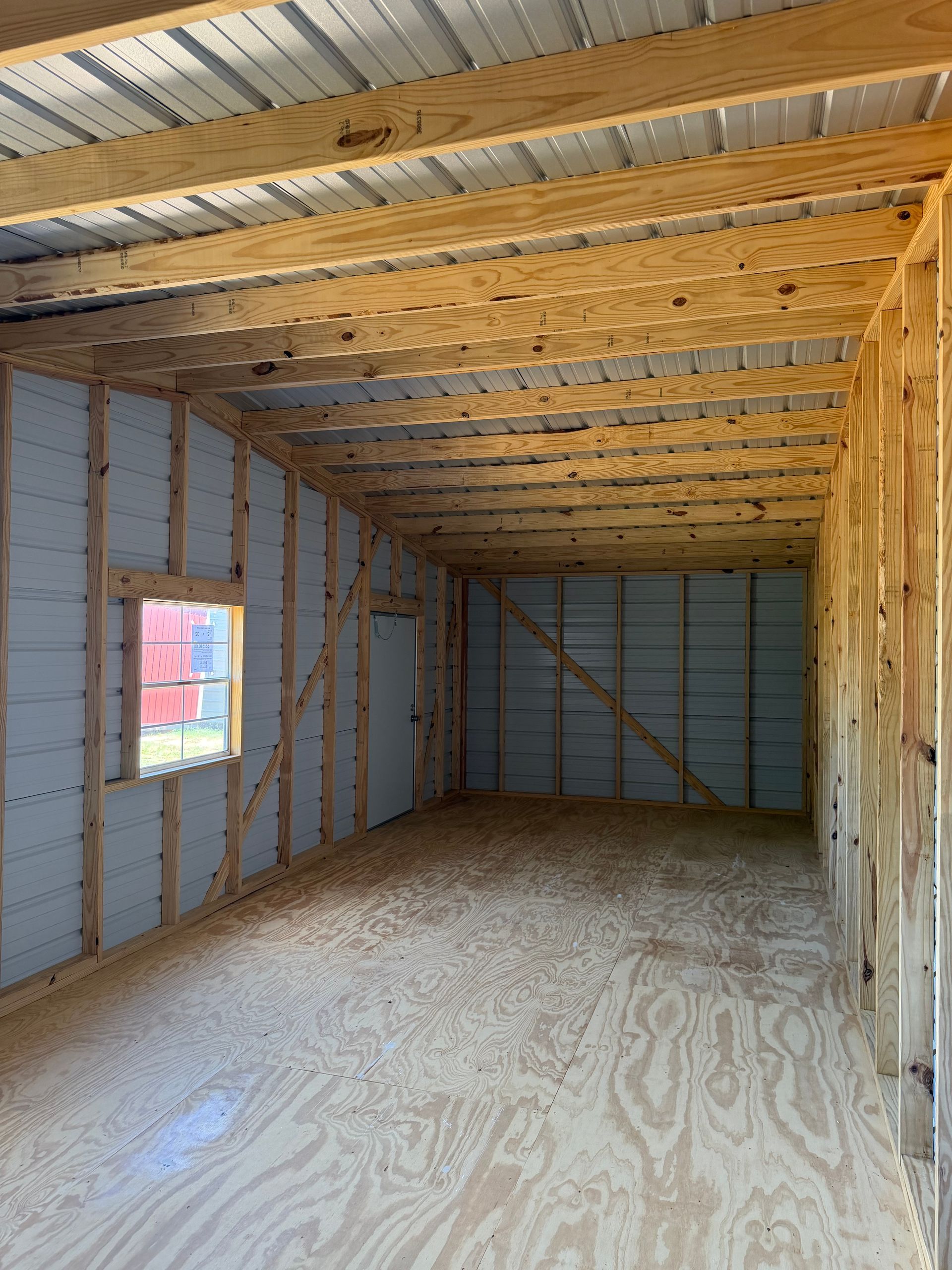 Empty unfinished room with plywood floor, exposed insulation, and a small window in the wall.