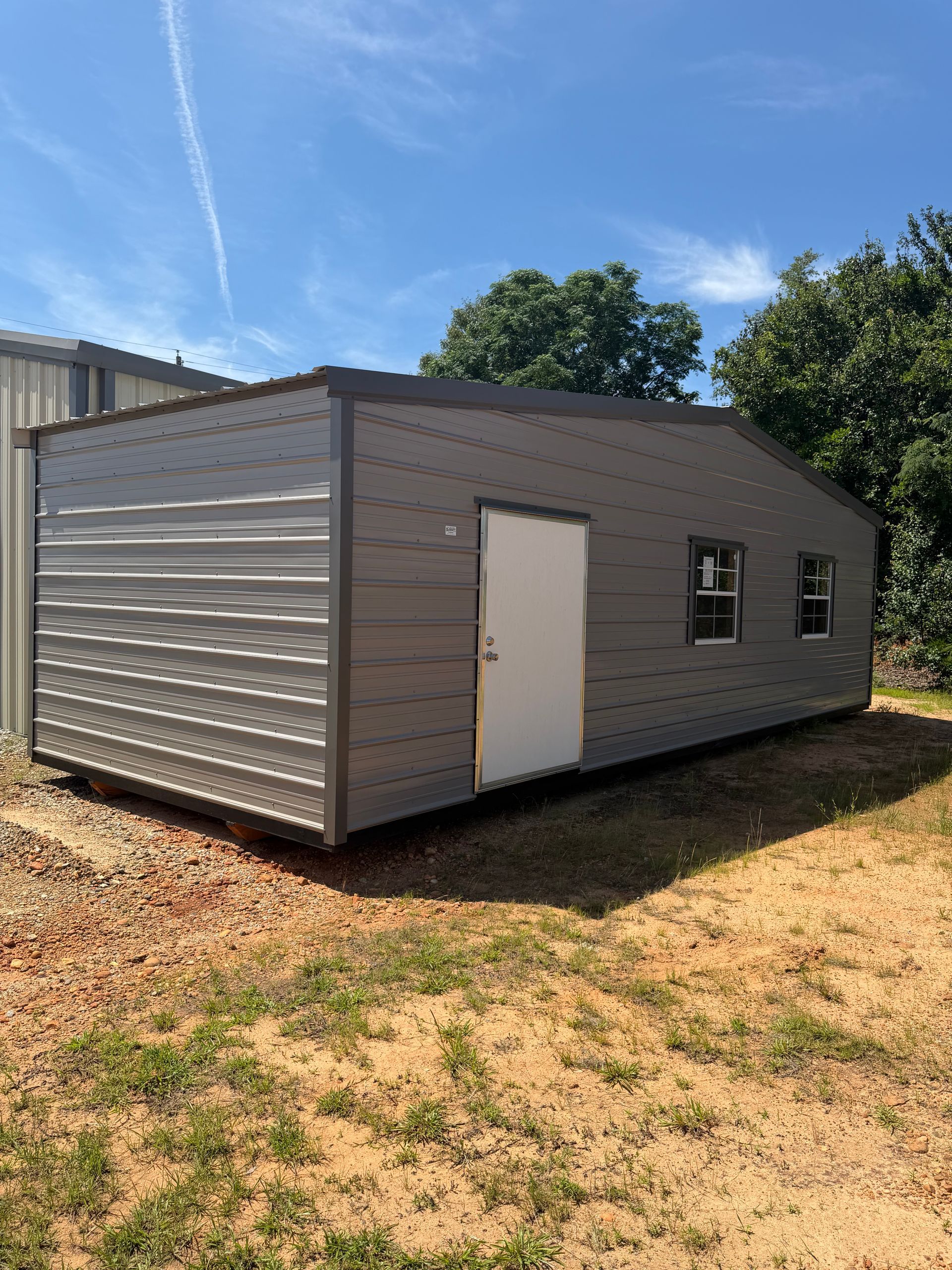 Gray metal storage shed with white door and small window on a dirt lot under a blue sky