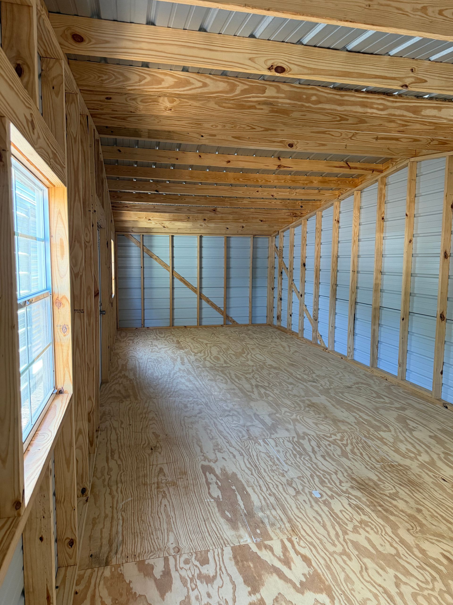 Empty unfinished wooden room with plywood walls, exposed ceiling joists, and a window on the left.