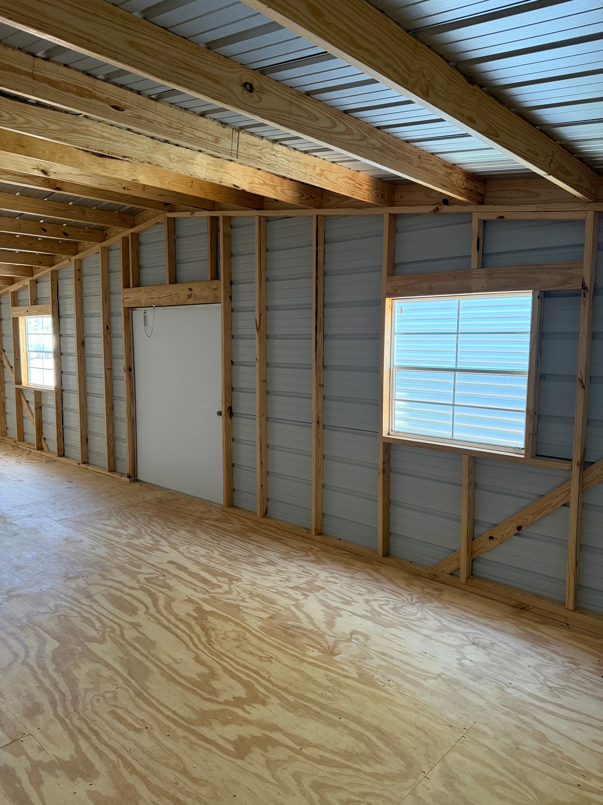 Unfinished room with exposed studs, plywood floor, and two windows in a partially insulated wall.