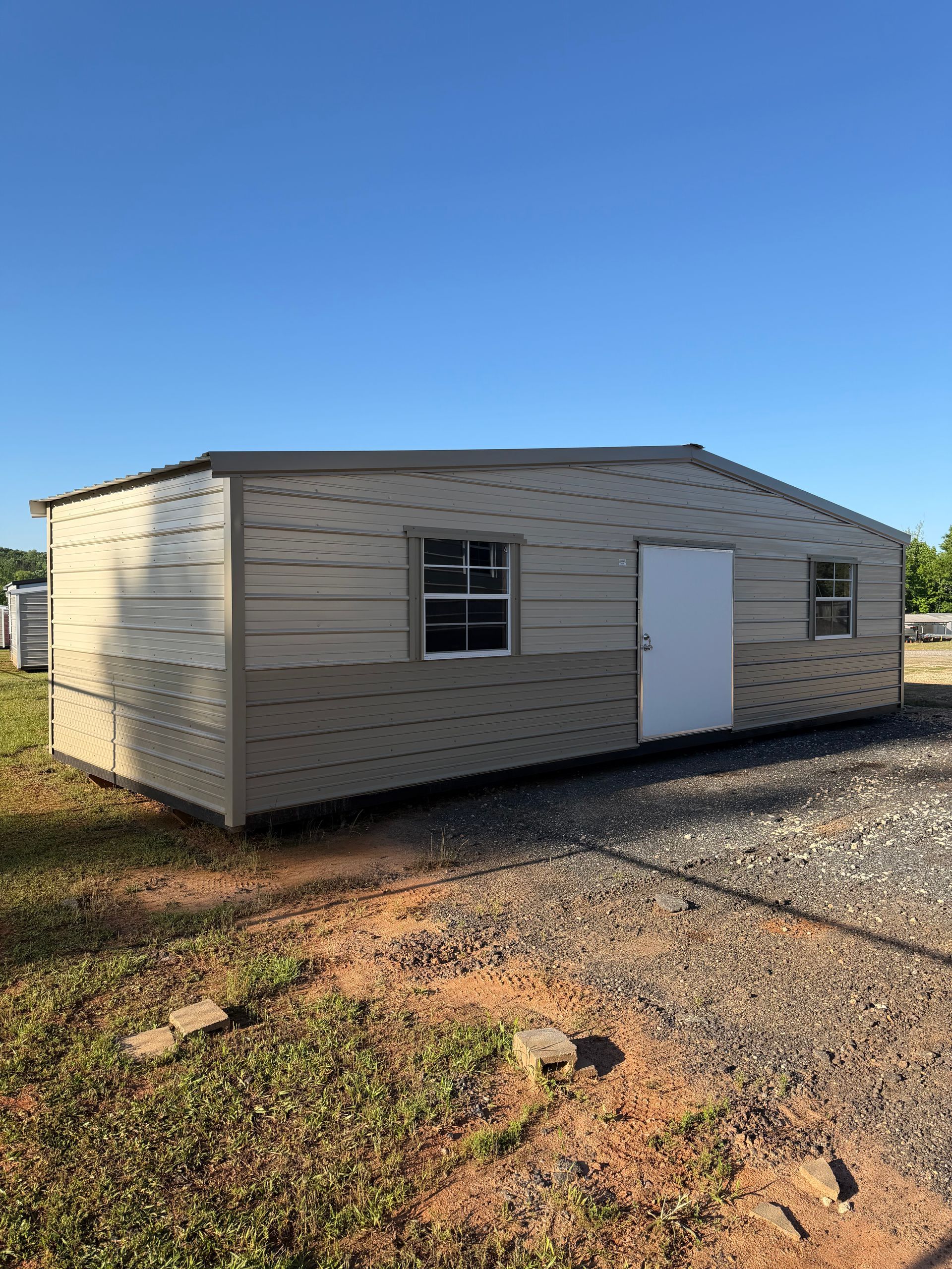 Small beige utility building with white door and windows on gravel under a clear blue sky