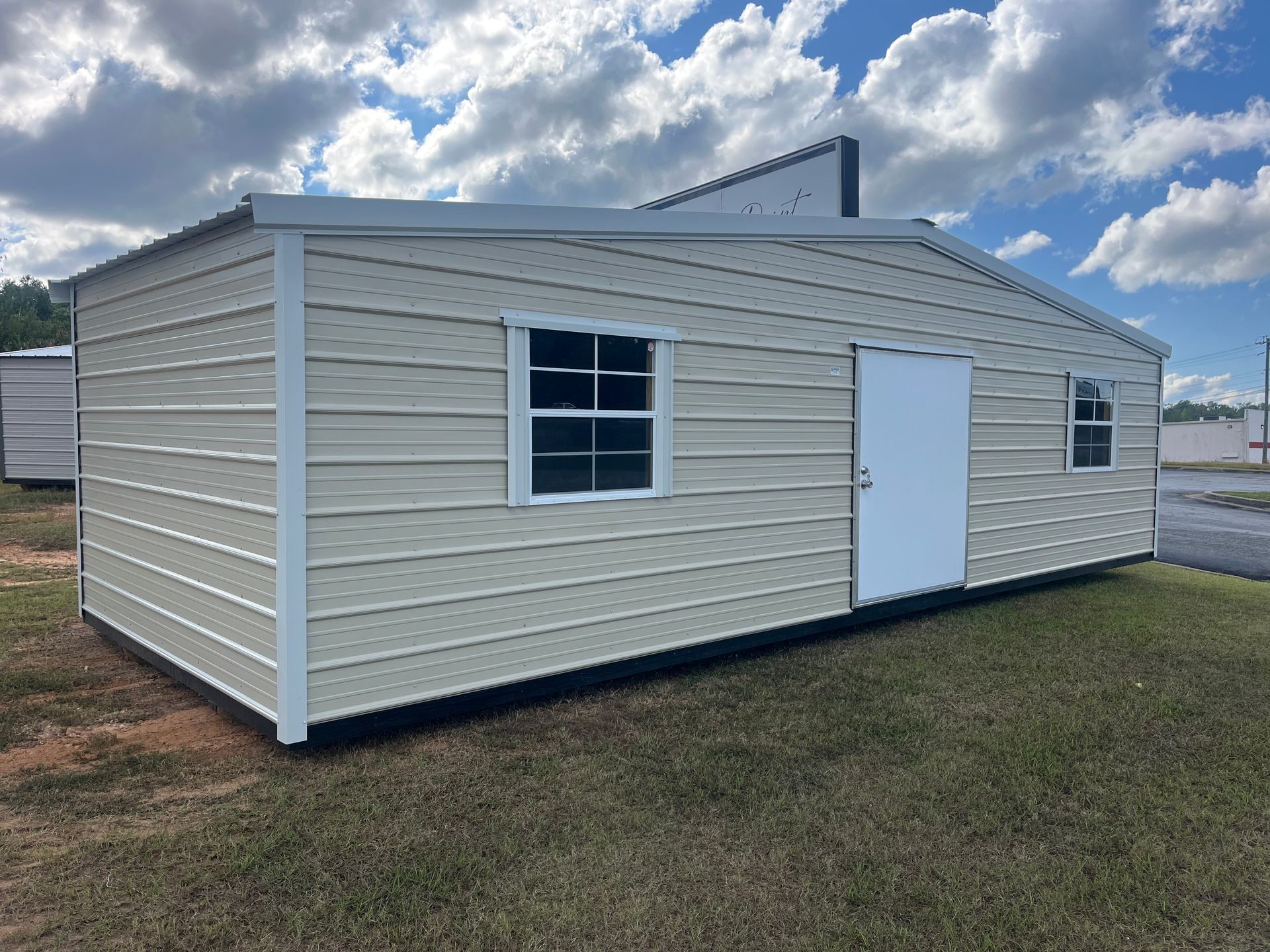 Beige metal shed with white door and window, standing on grass under cloudy sky