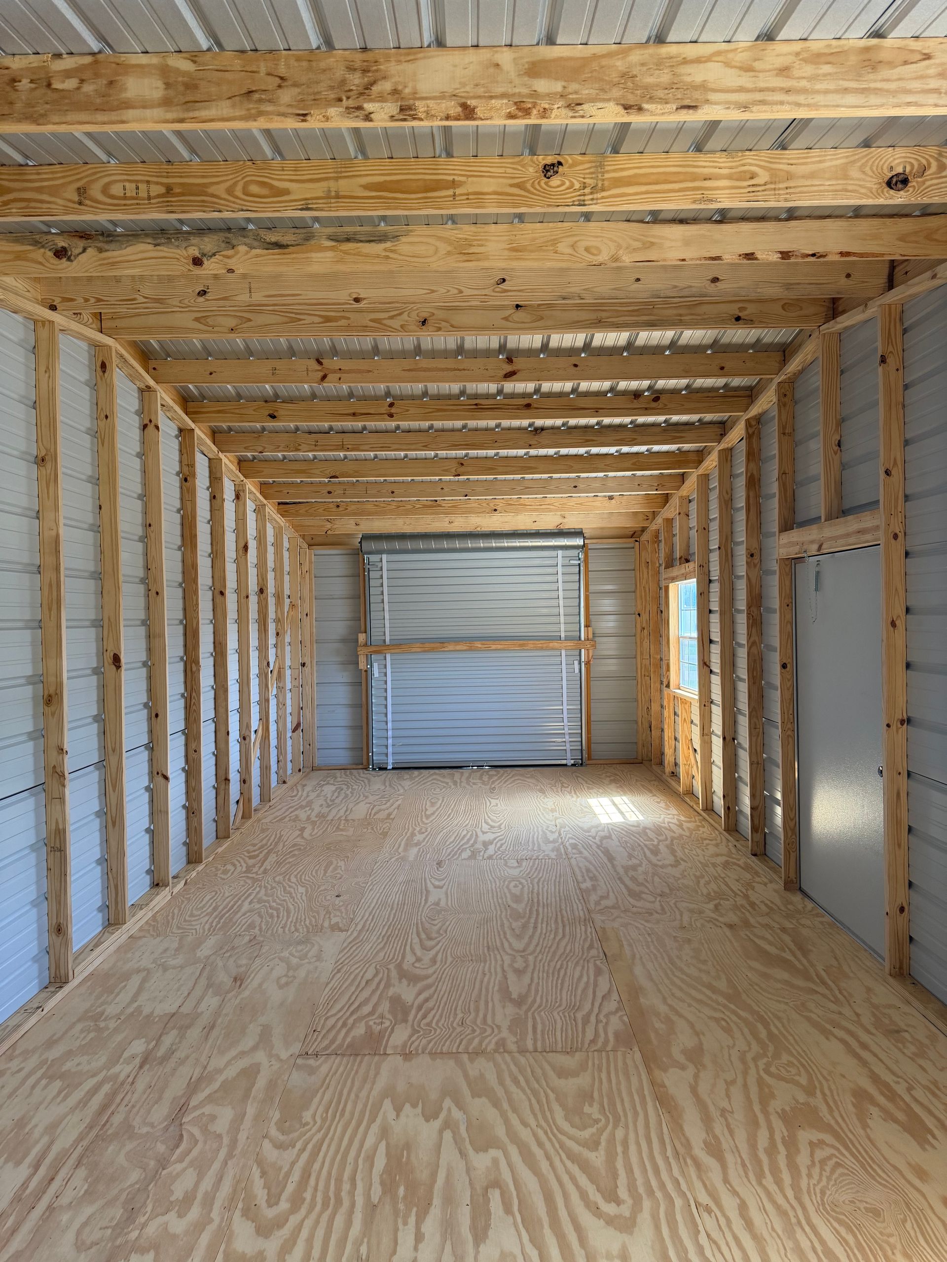 Empty unfinished room with exposed wood framing, plywood floor, and a closed garage door at the far end