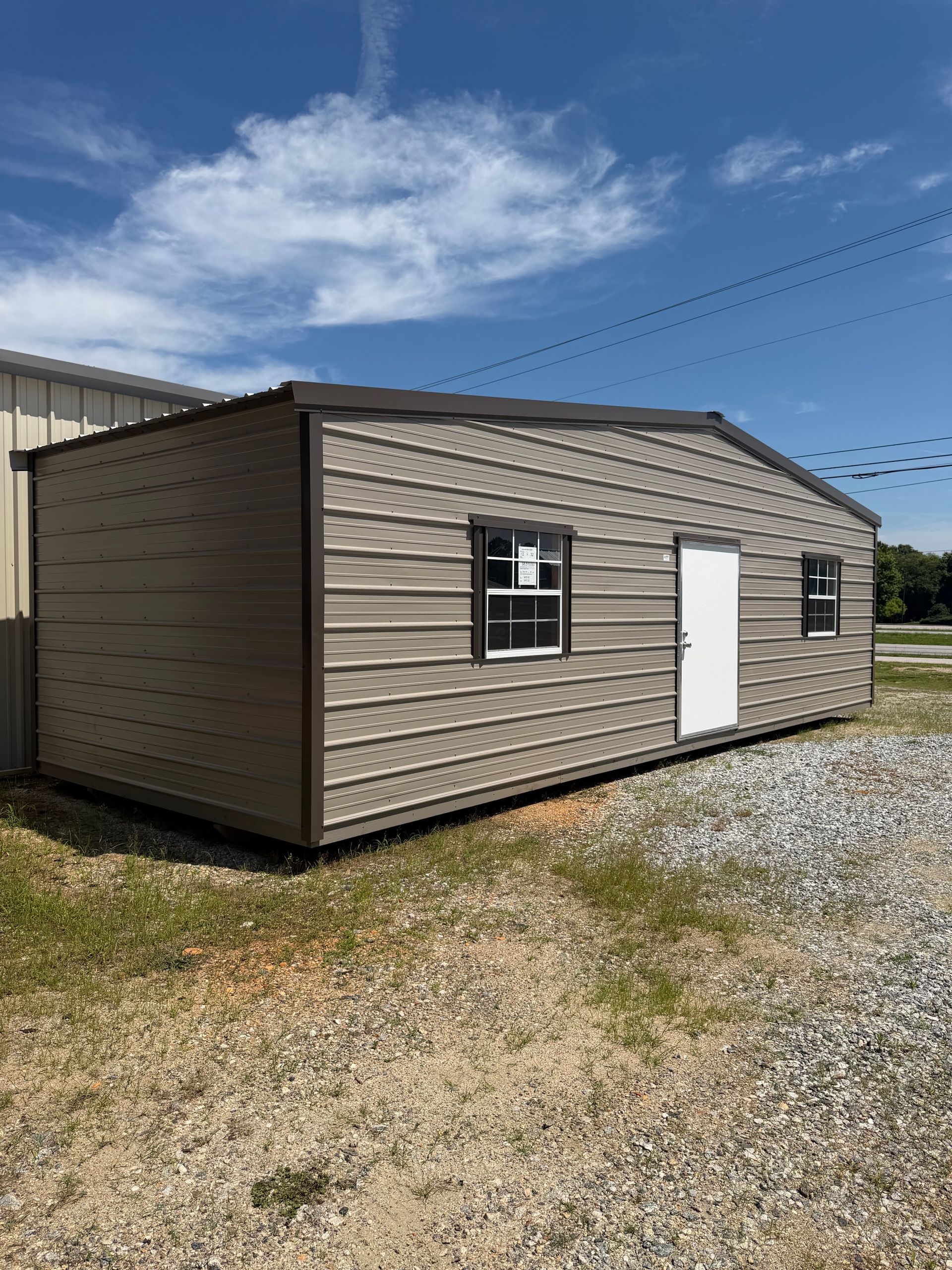 Small tan storage building with white door and black window on gravel under a blue sky.