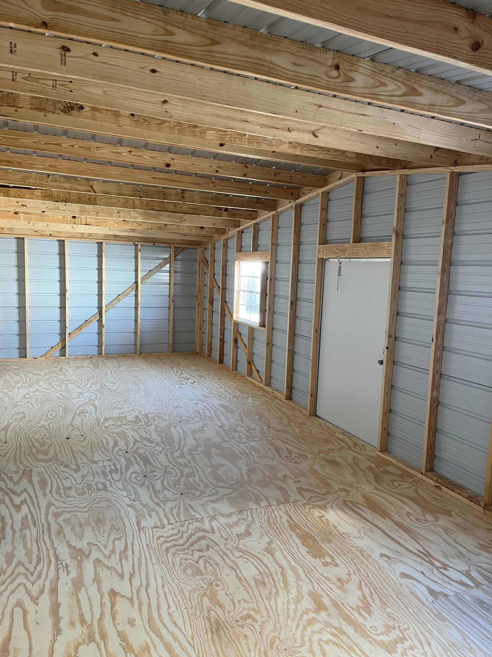 Unfinished attic with exposed studs, plywood floor, and a small window and door opening along the wall