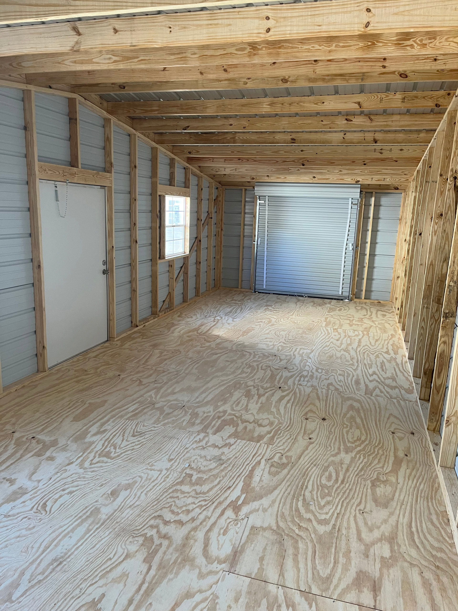 Unfinished room with exposed studs, plywood floor, and taped-off opening at the far end.