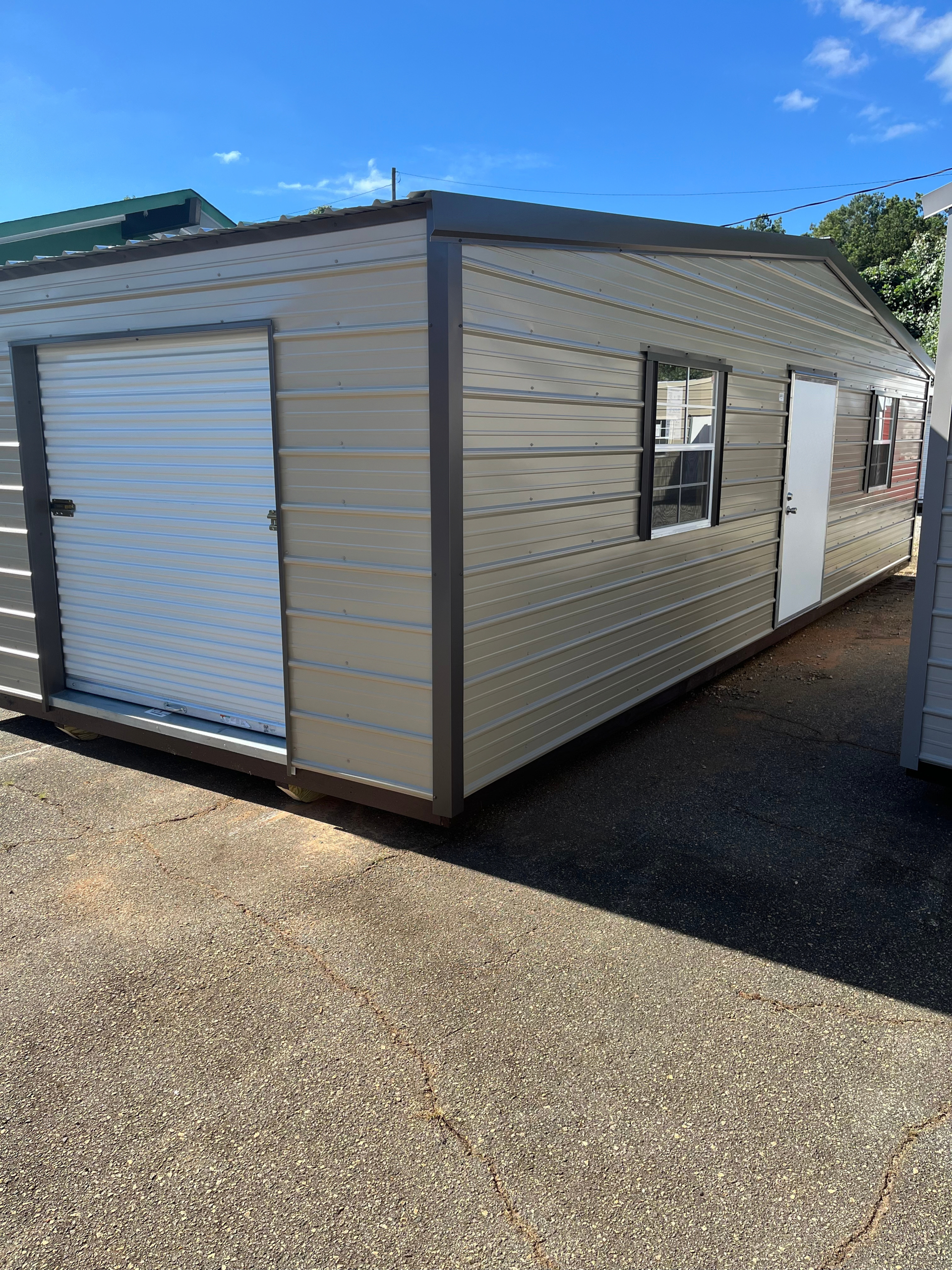 Small beige metal shed with white roll-up door and window on a gravel lot under a blue sky
