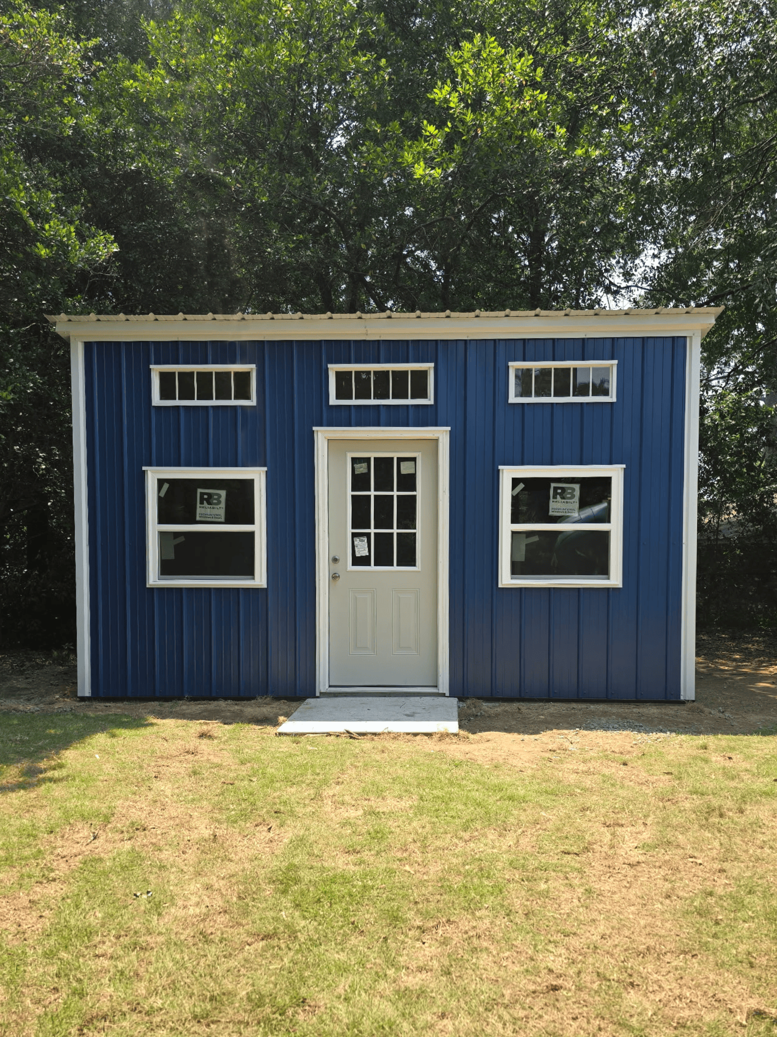 Small blue shed with white trim, front door, and windows in a grassy yard with trees behind it