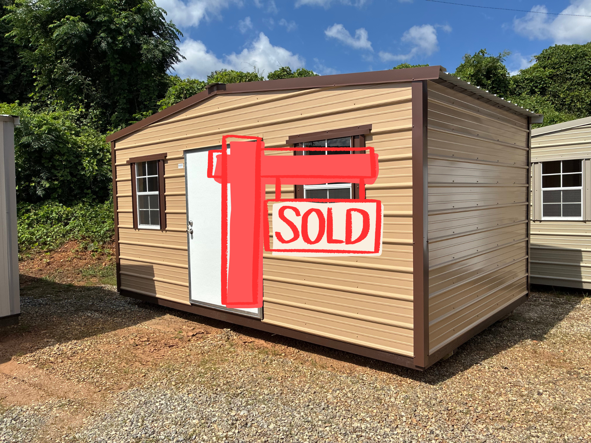 Small tan shed with a red “SOLD” sign outside on a gravel lot.