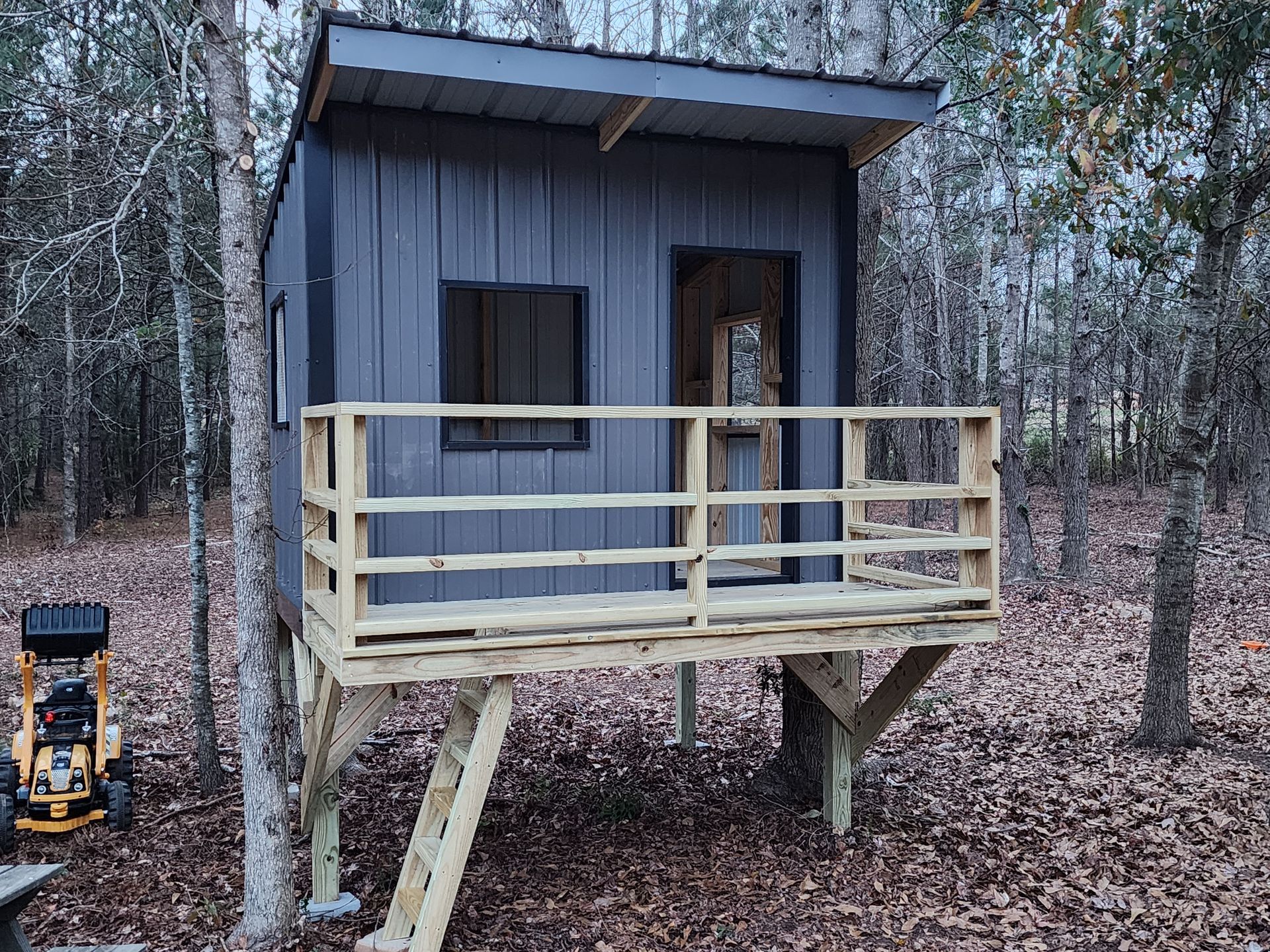 Small gray hunting cabin on stilts with a wooden deck in a leaf-covered forest