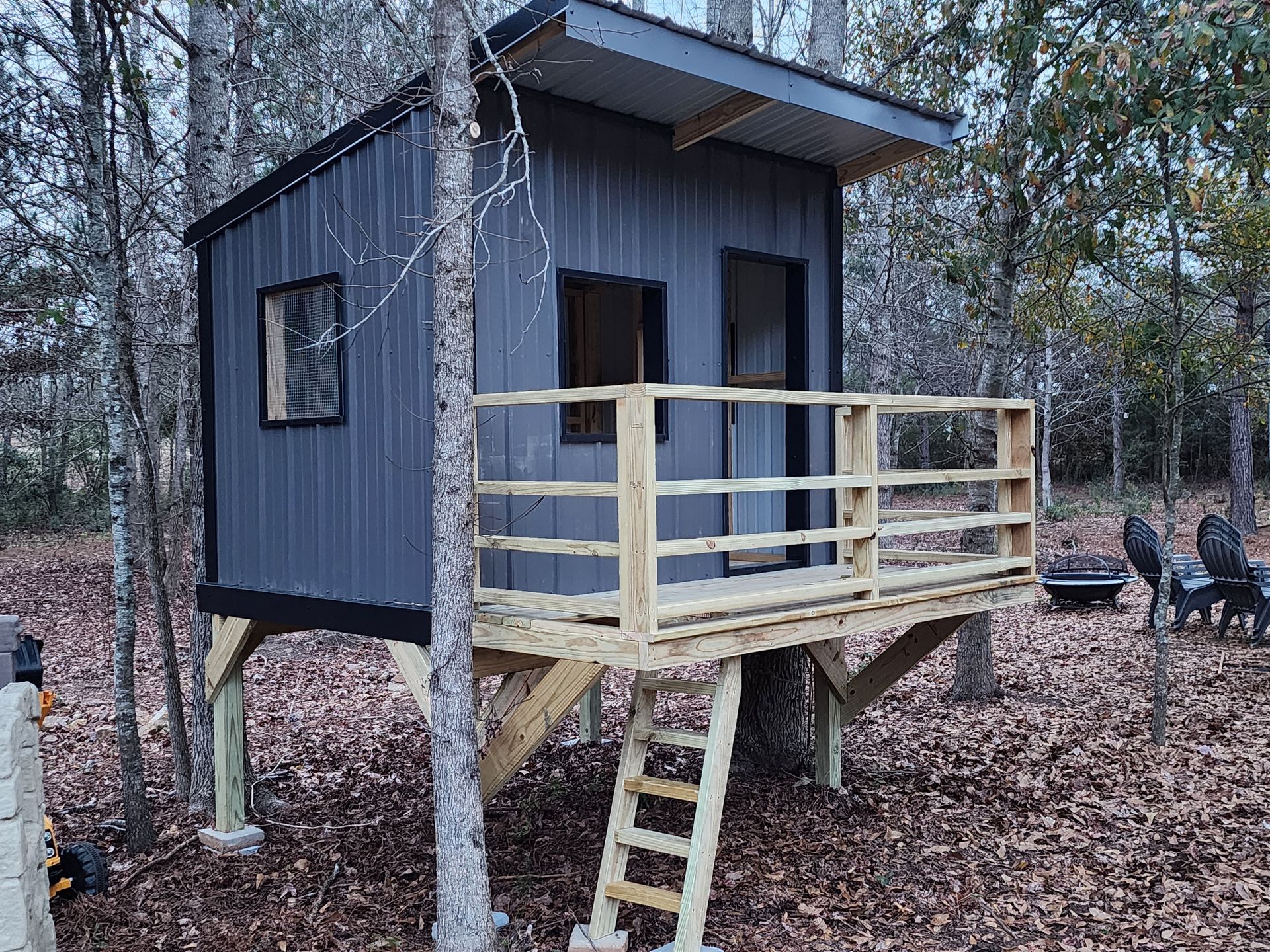 Small gray cabin on stilts with a porch and ladder in a wooded area