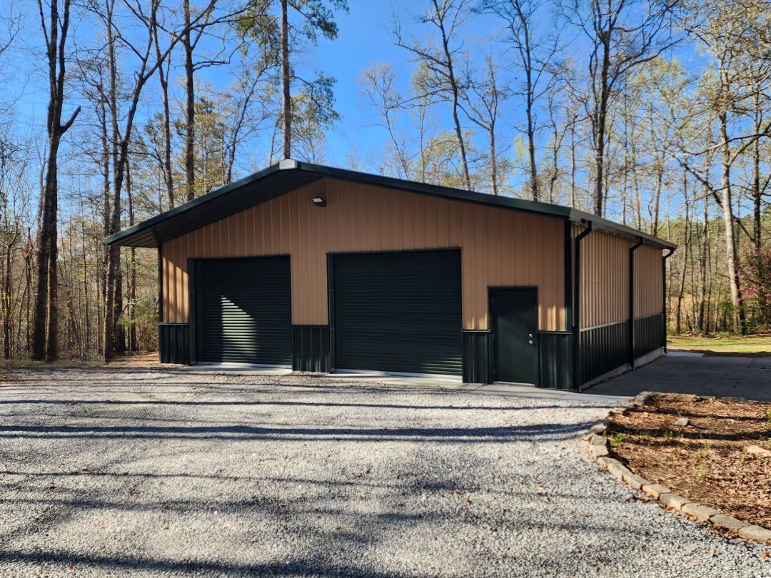 Two-bay tan and green metal garage in a wooded area with gravel driveway.