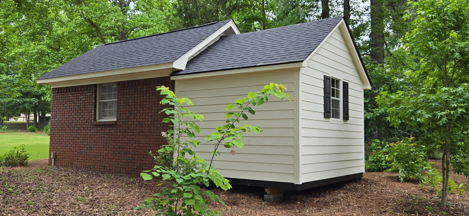 Small rustic shed with red brick and white siding in a wooded yard