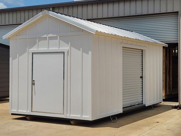White storage shed with a roll-up door and a single door, set on a concrete pad.