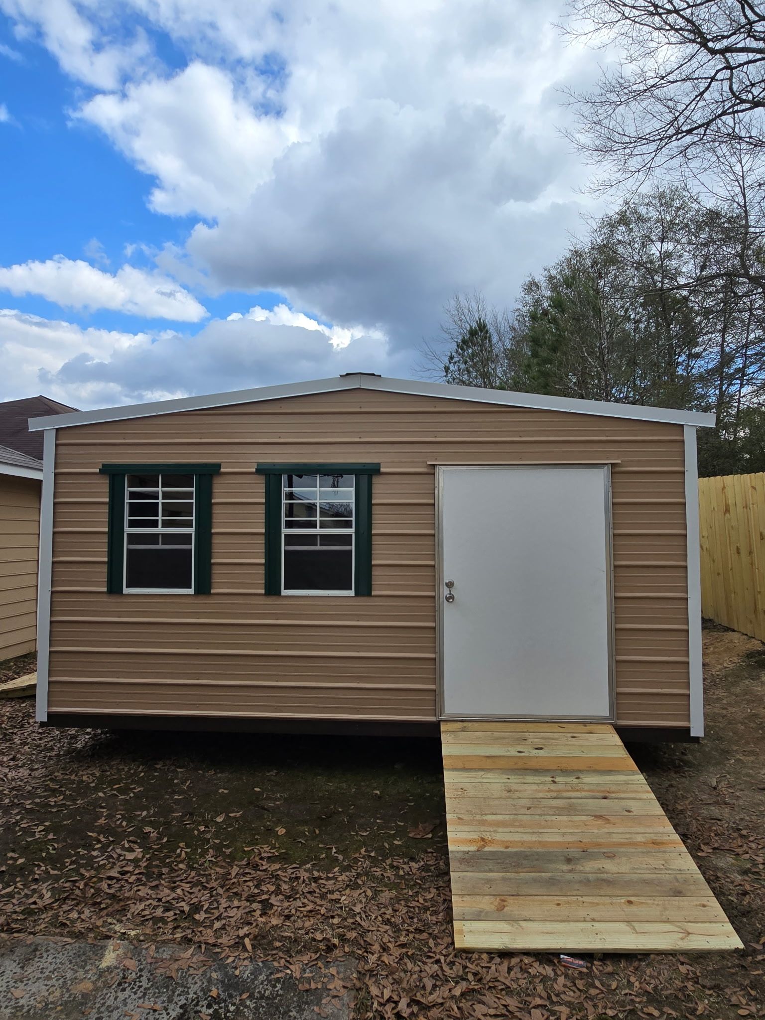 Tan shed with a white door, two windows, and a wooden ramp. Green trim on the windows. Cloudy sky.