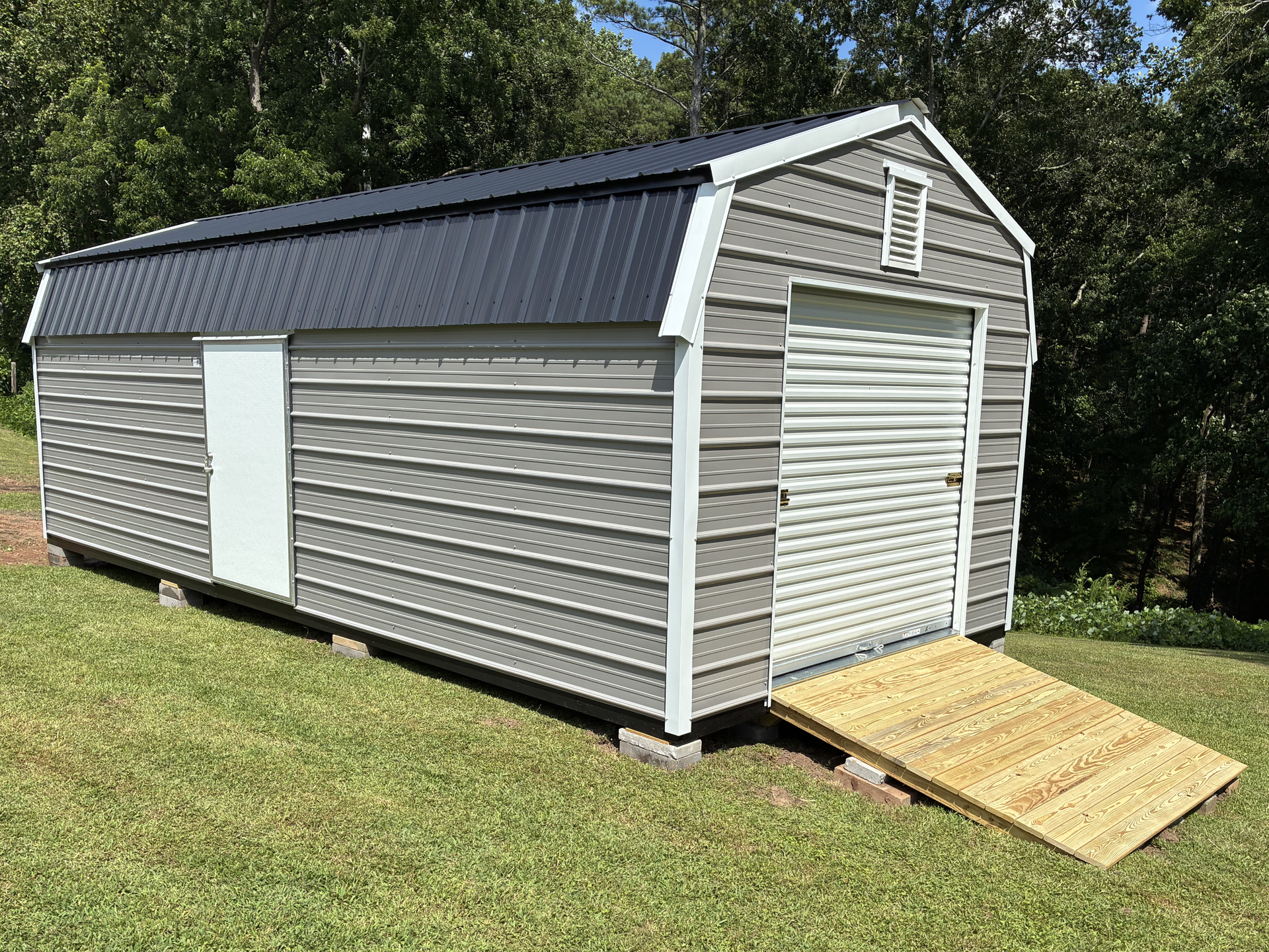 White shed with gray siding and black roof on a grassy lawn, with a ramp at the front.