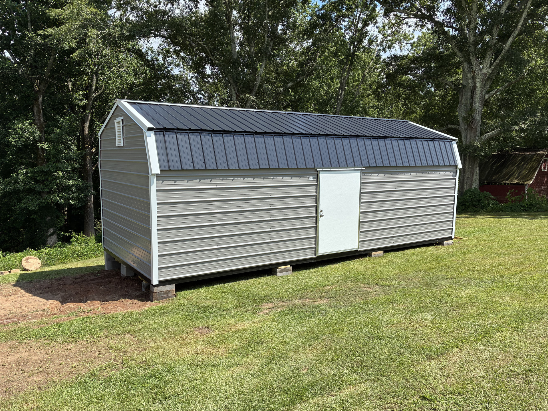 Gray shed with dark roof and white door on a grassy lawn by trees