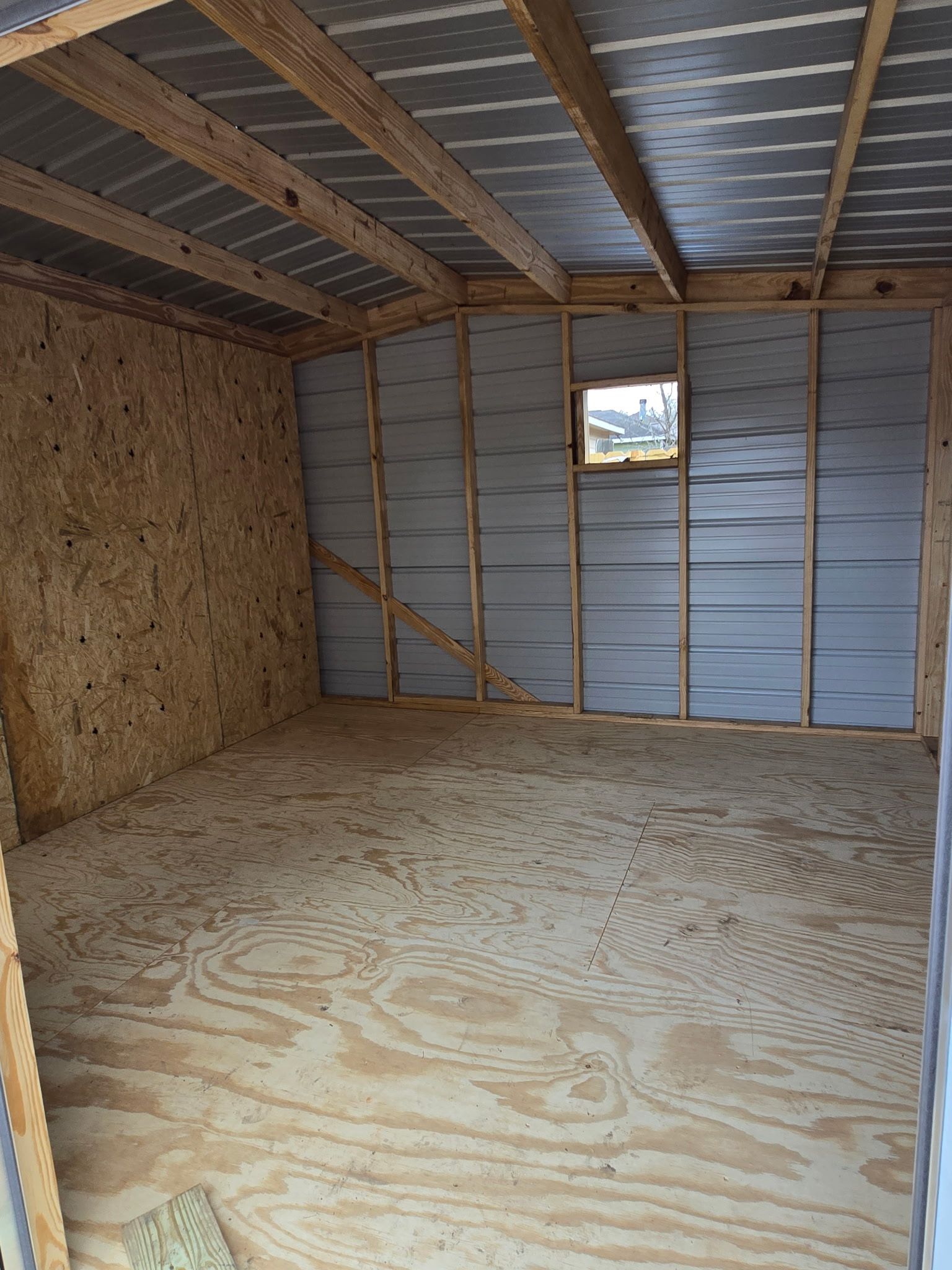 Interior view of a partially built shed. Walls and floor made of wood; metal roofing and siding visible. Small window.