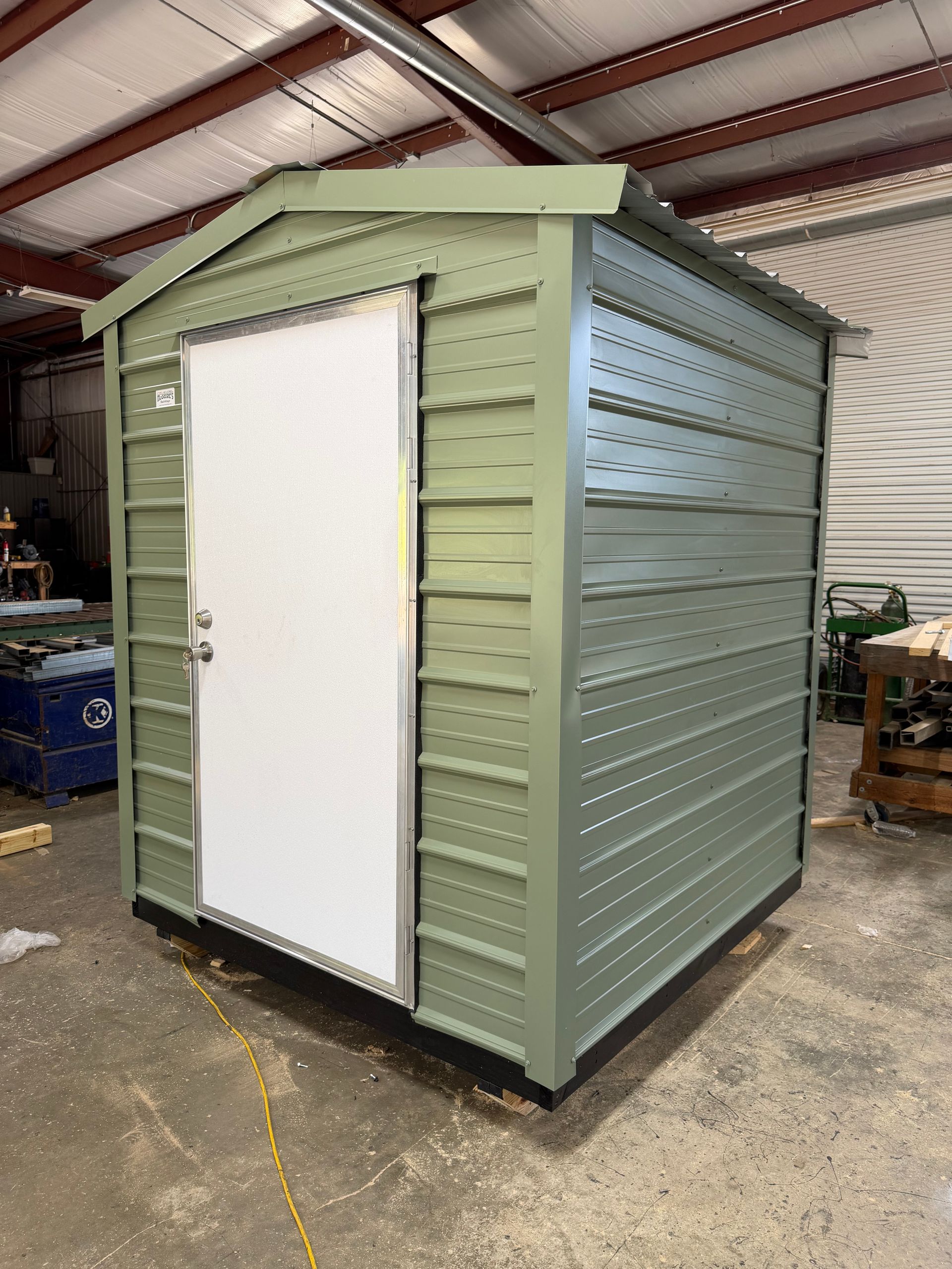Small green metal shed with a white door inside a warehouse-like workshop.