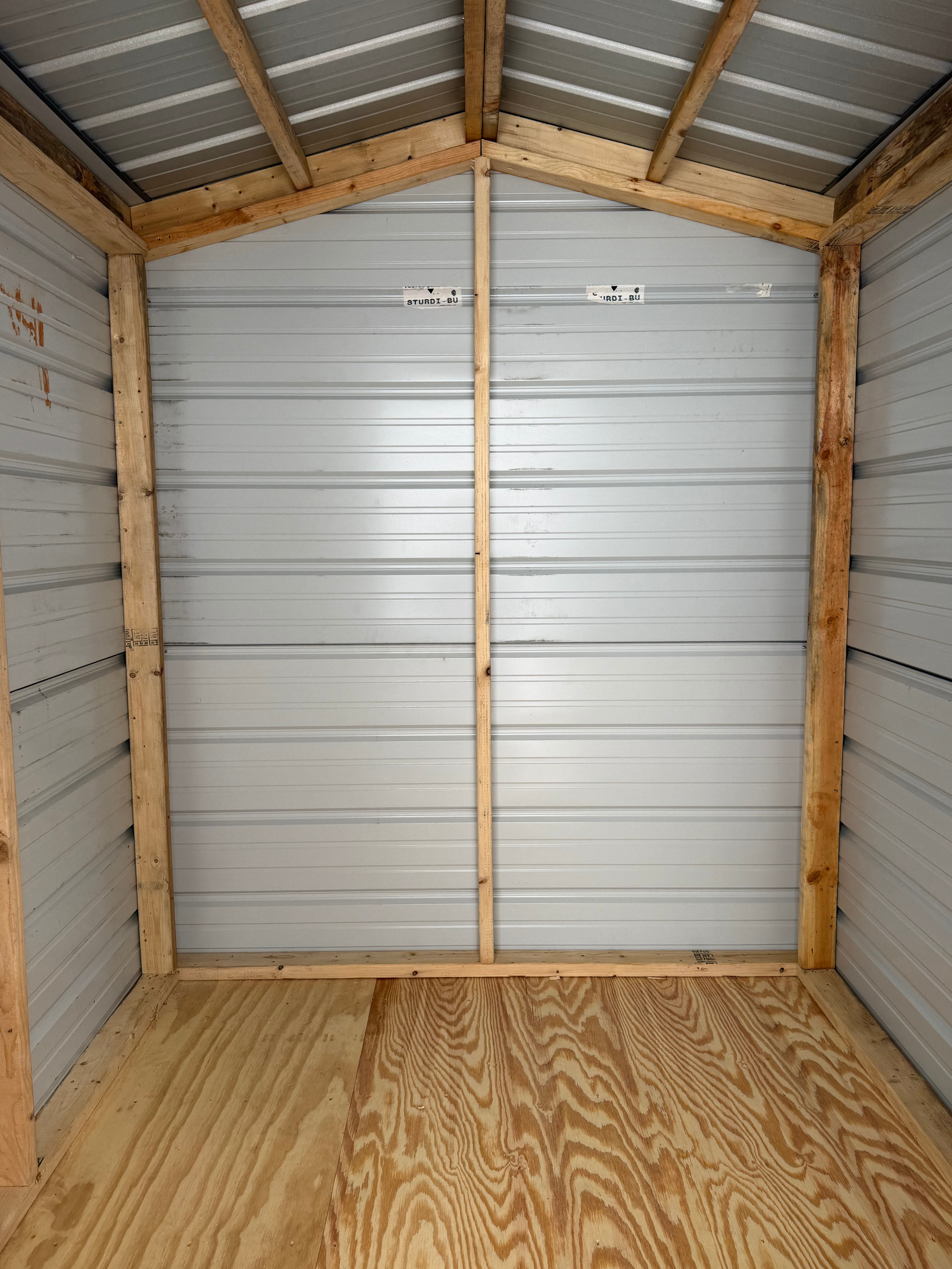 Empty storage shed with plywood floor, white metal walls, and corrugated metal roof panels