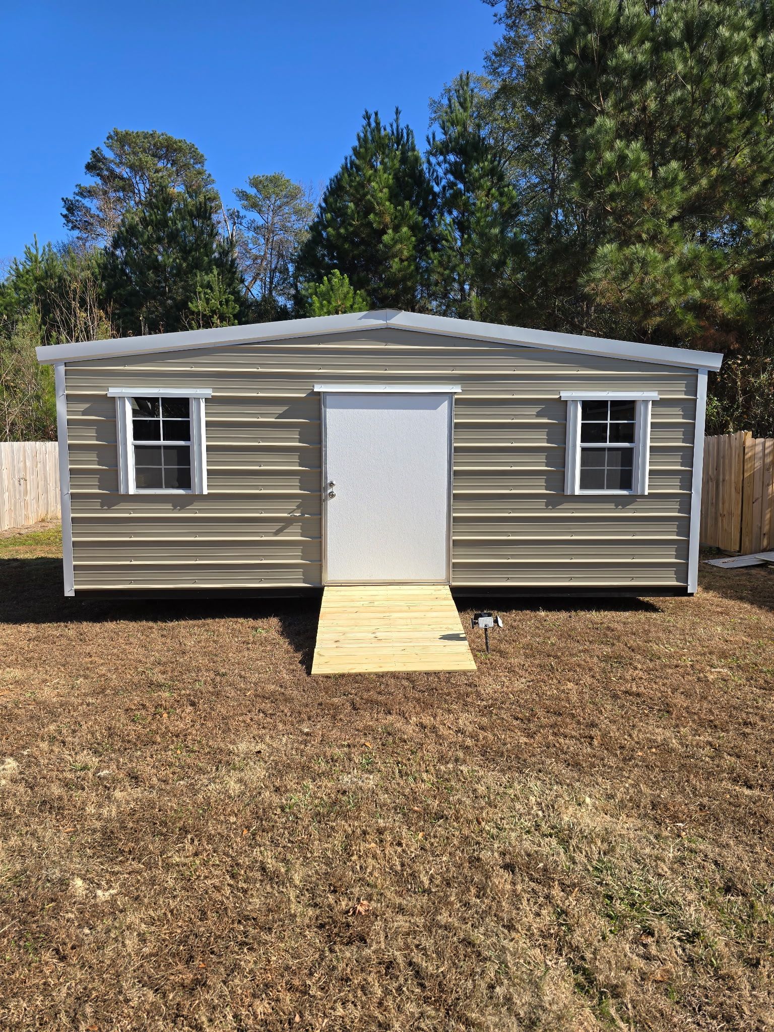 Metal shed with door and two windows, brown siding, wooden ramp, set on brown grass, trees in background.