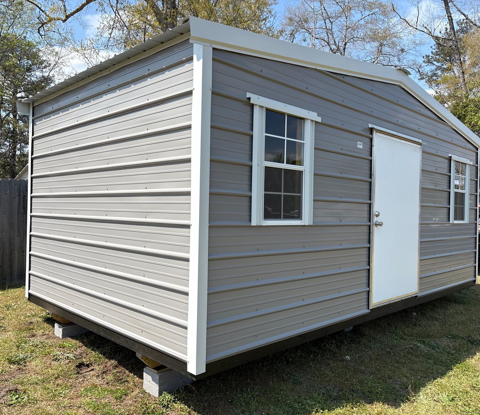 A gray metal shed with white trim and a white door, elevated on concrete blocks in a grassy area with trees behind.