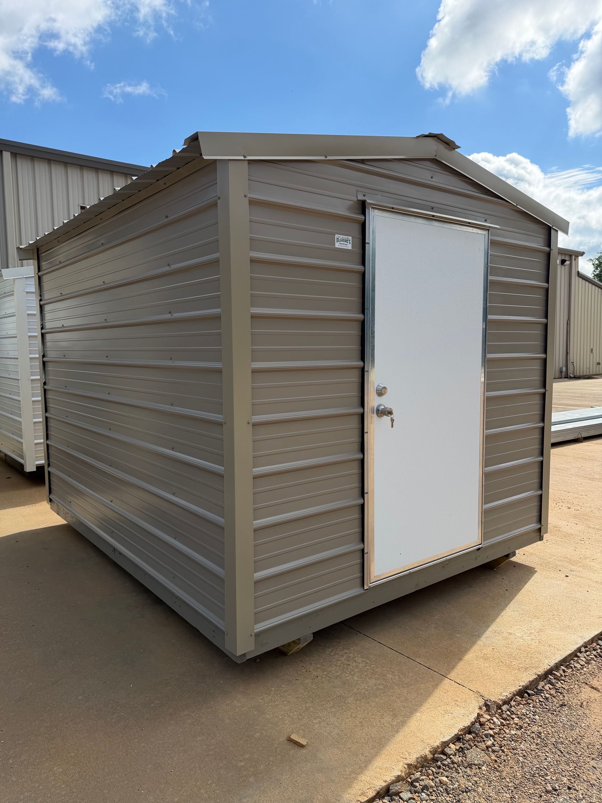Small gray metal storage shed with a white door on a paved outdoor lot.