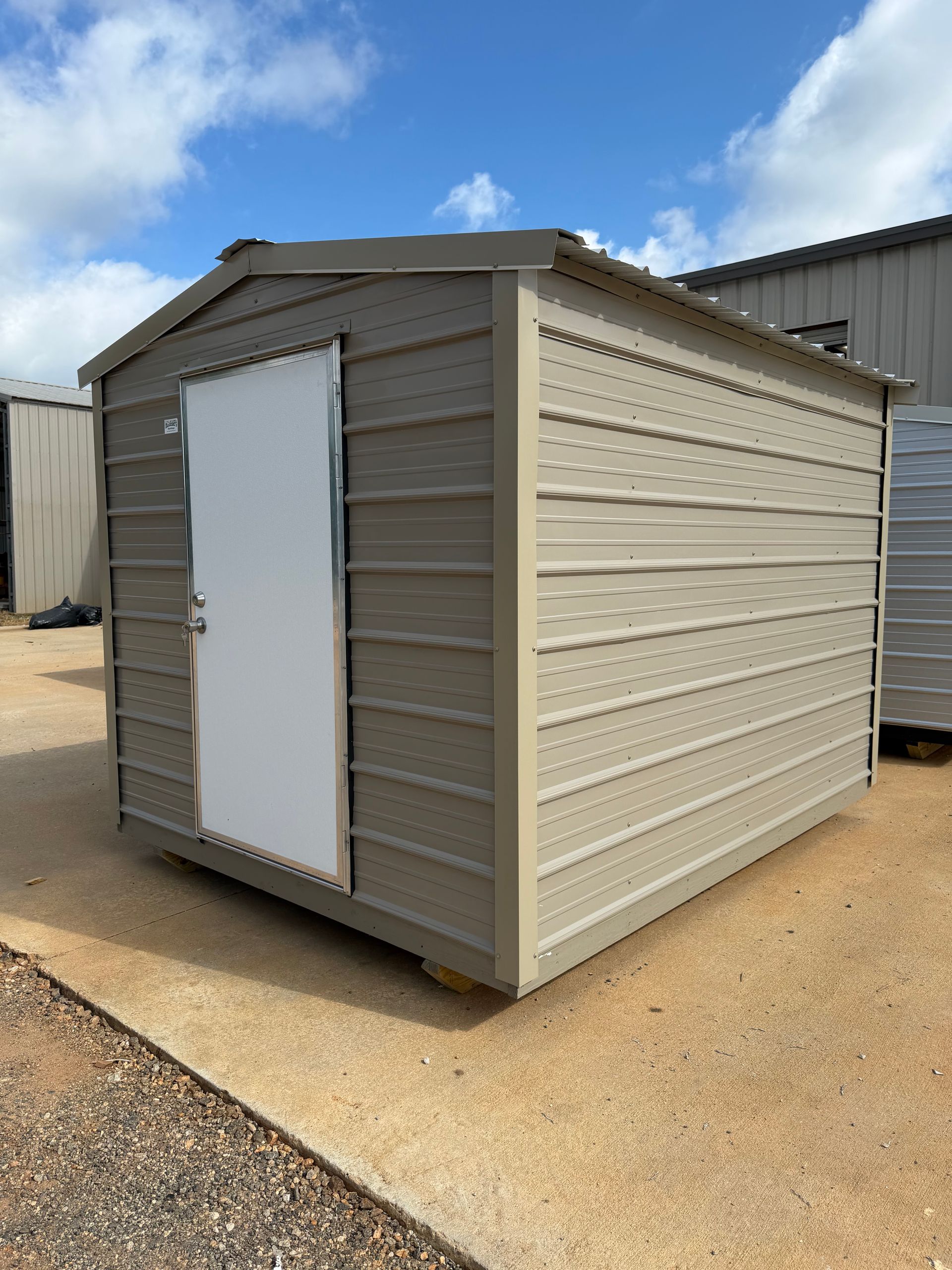 Small tan outdoor shed with a white door on gravel under a partly cloudy sky