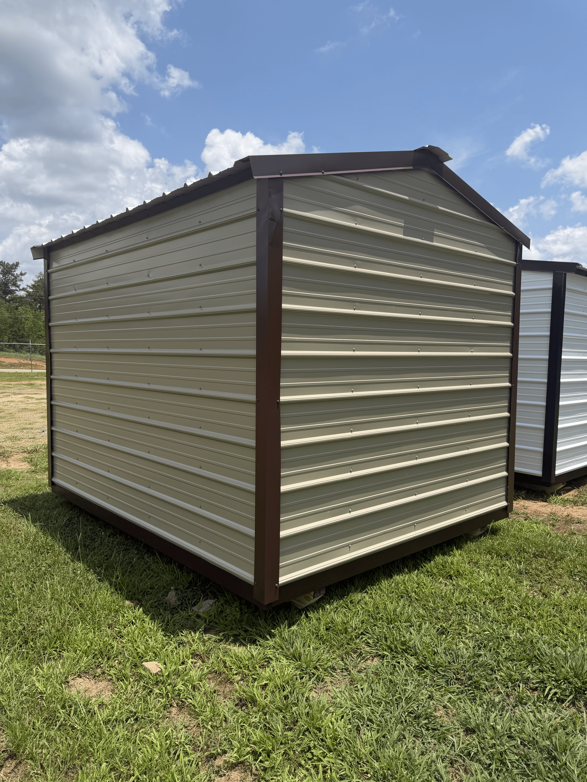 Beige corrugated metal storage shed with brown trim on grass under a blue sky