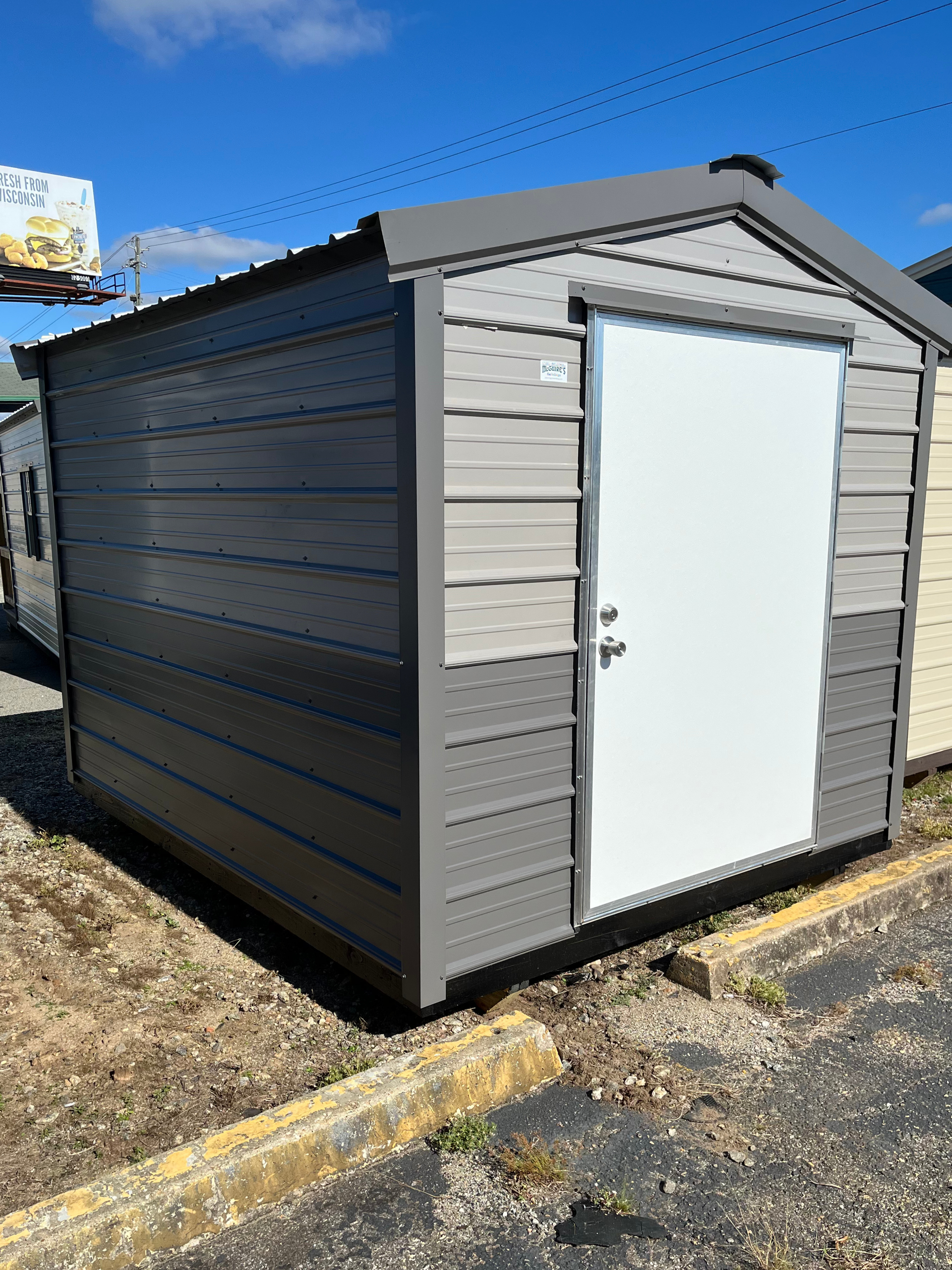 Small gray storage shed with a white door beside a curb on a sunny day