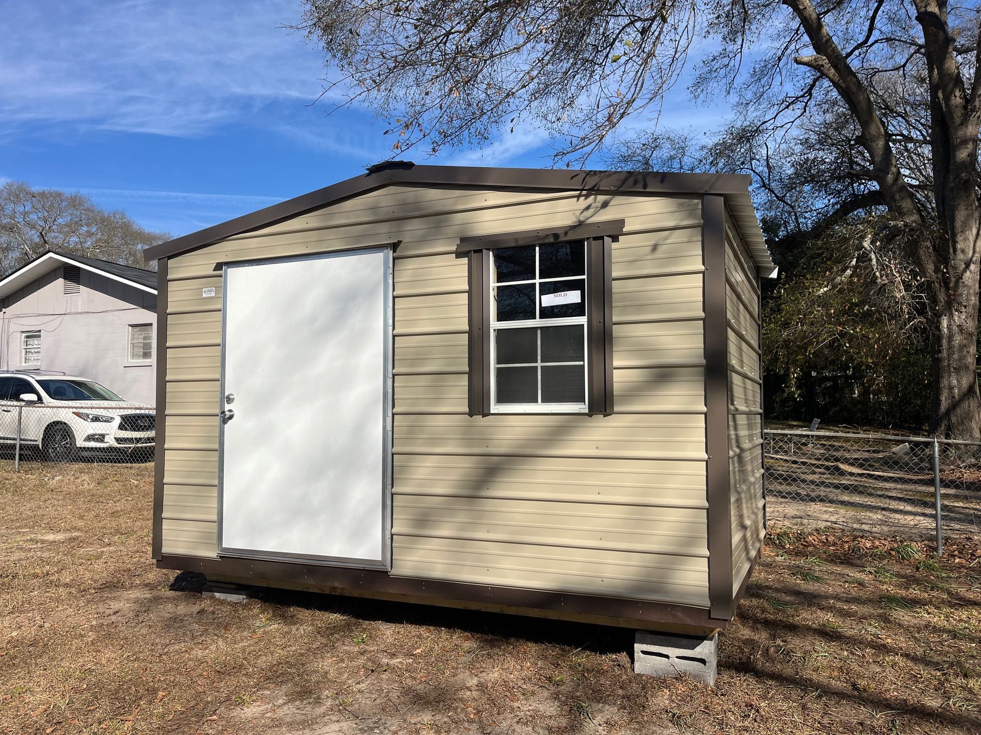 Tan shed with white door and window, brown trim, sitting on cinder blocks in a yard.