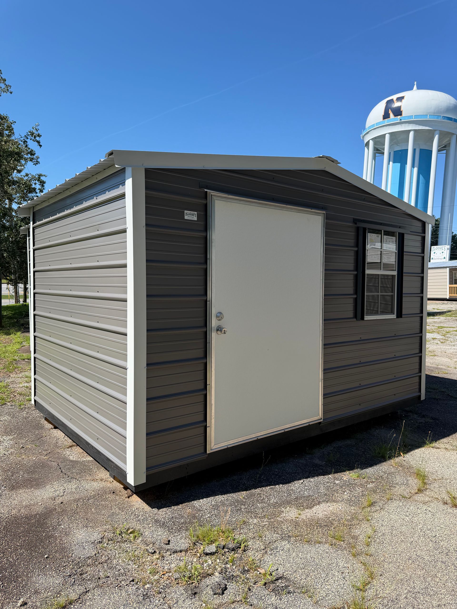 Small gray shed with white door on gravel under a clear blue sky, with a water tower in the background.