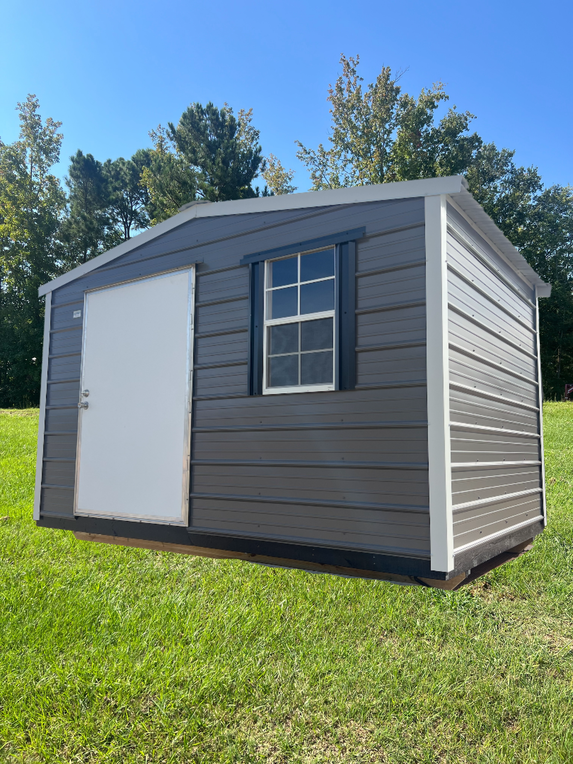Small gray shed with white trim, a door and window, sitting on a grassy lawn under a blue sky.
