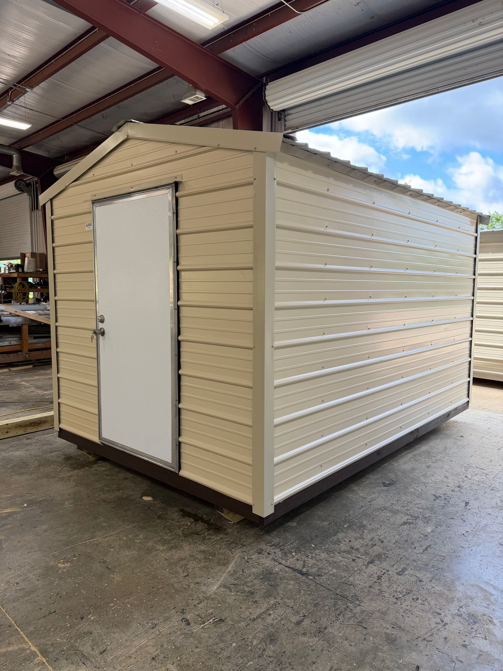 Beige storage shed with white door inside a metal-roofed warehouse.