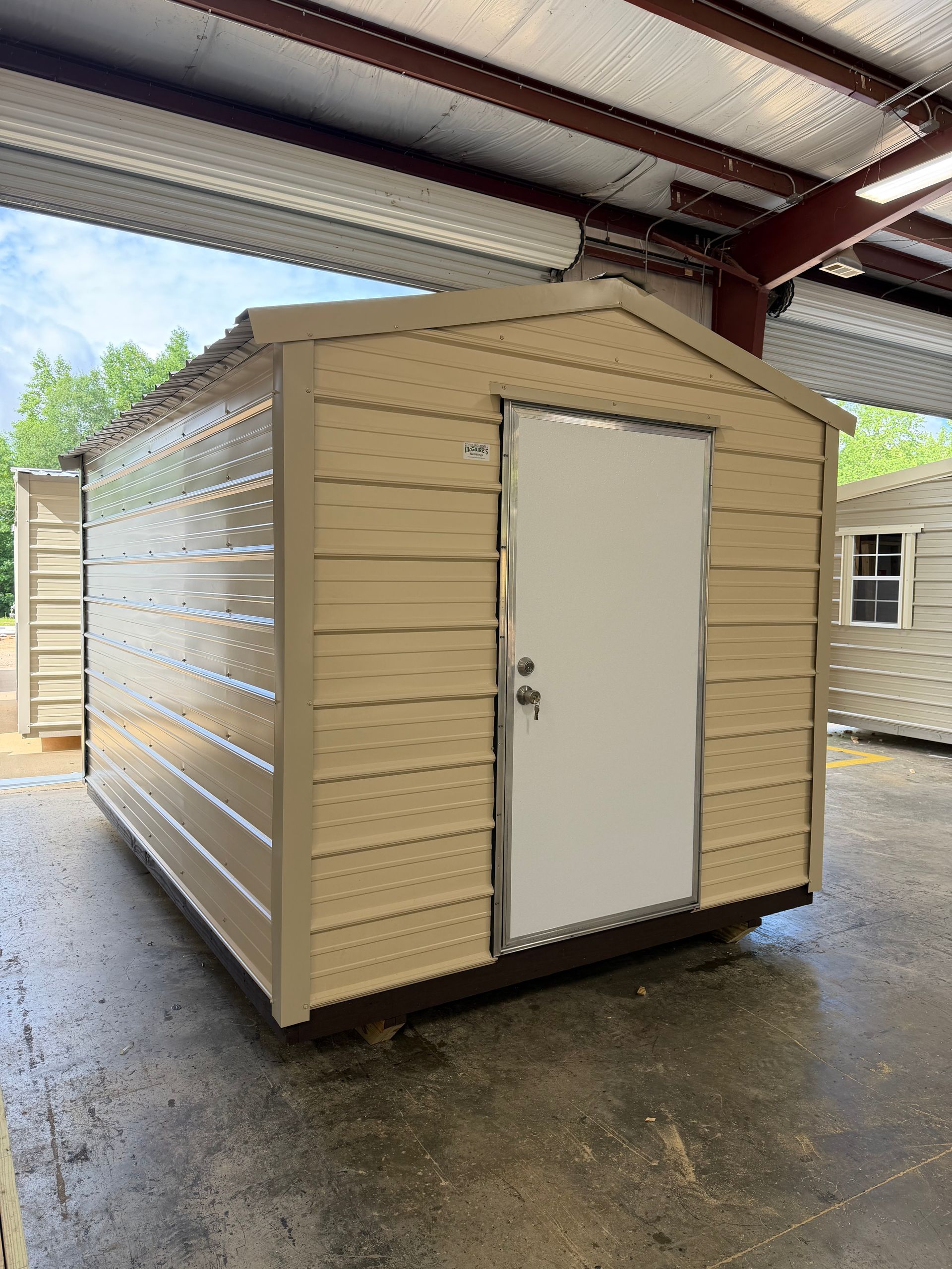 Beige storage shed with white door under a metal carport, wet floor beneath it.
