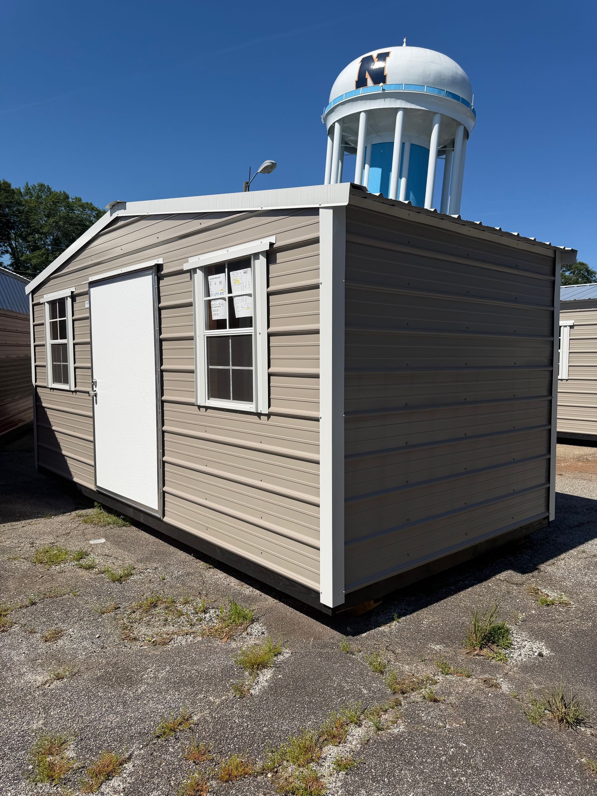 Small beige shed with white trim on a gravel lot, beside a large white water tower under a blue sky