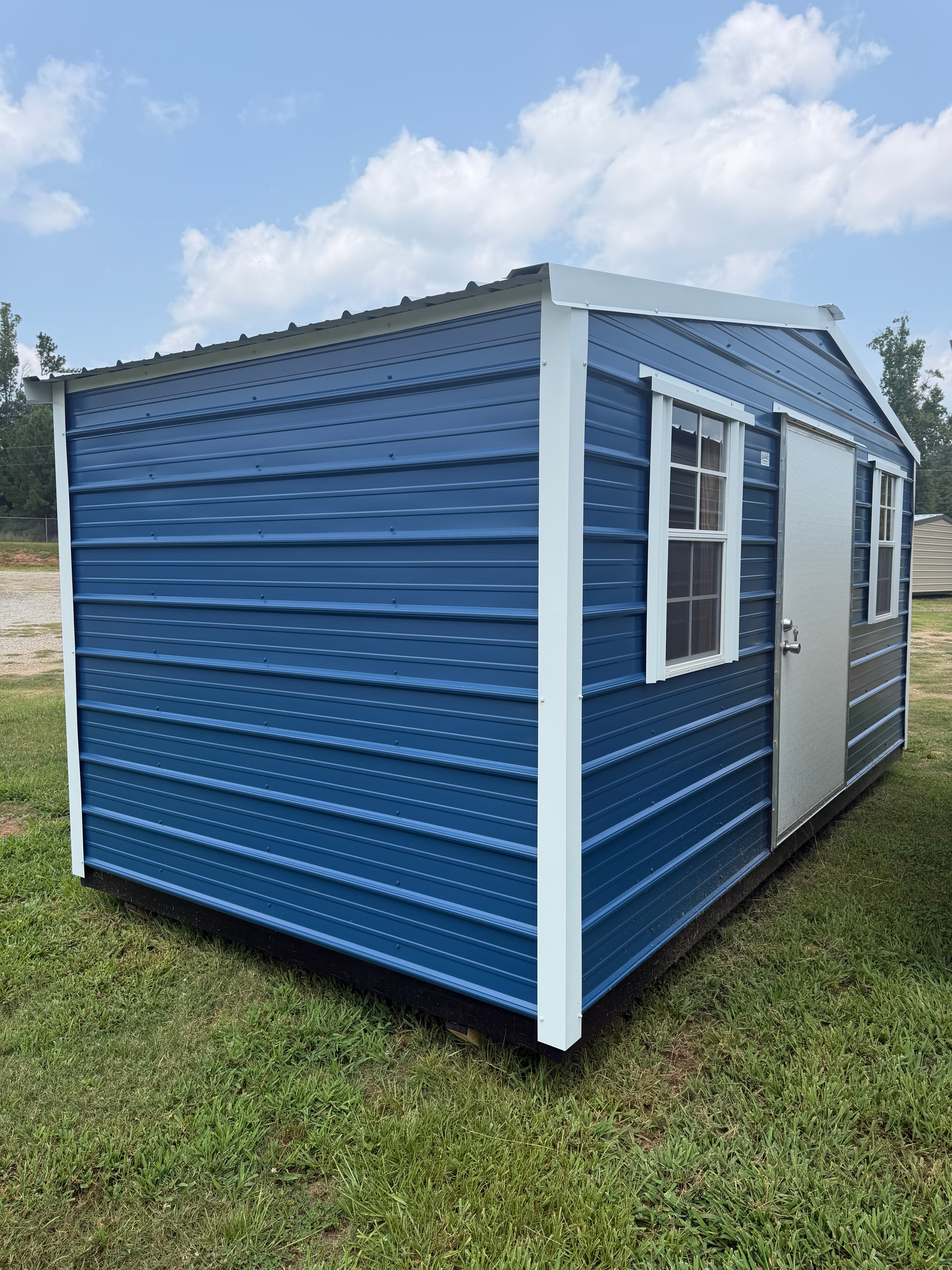 Blue portable shed with white trim and windows on a grassy lot