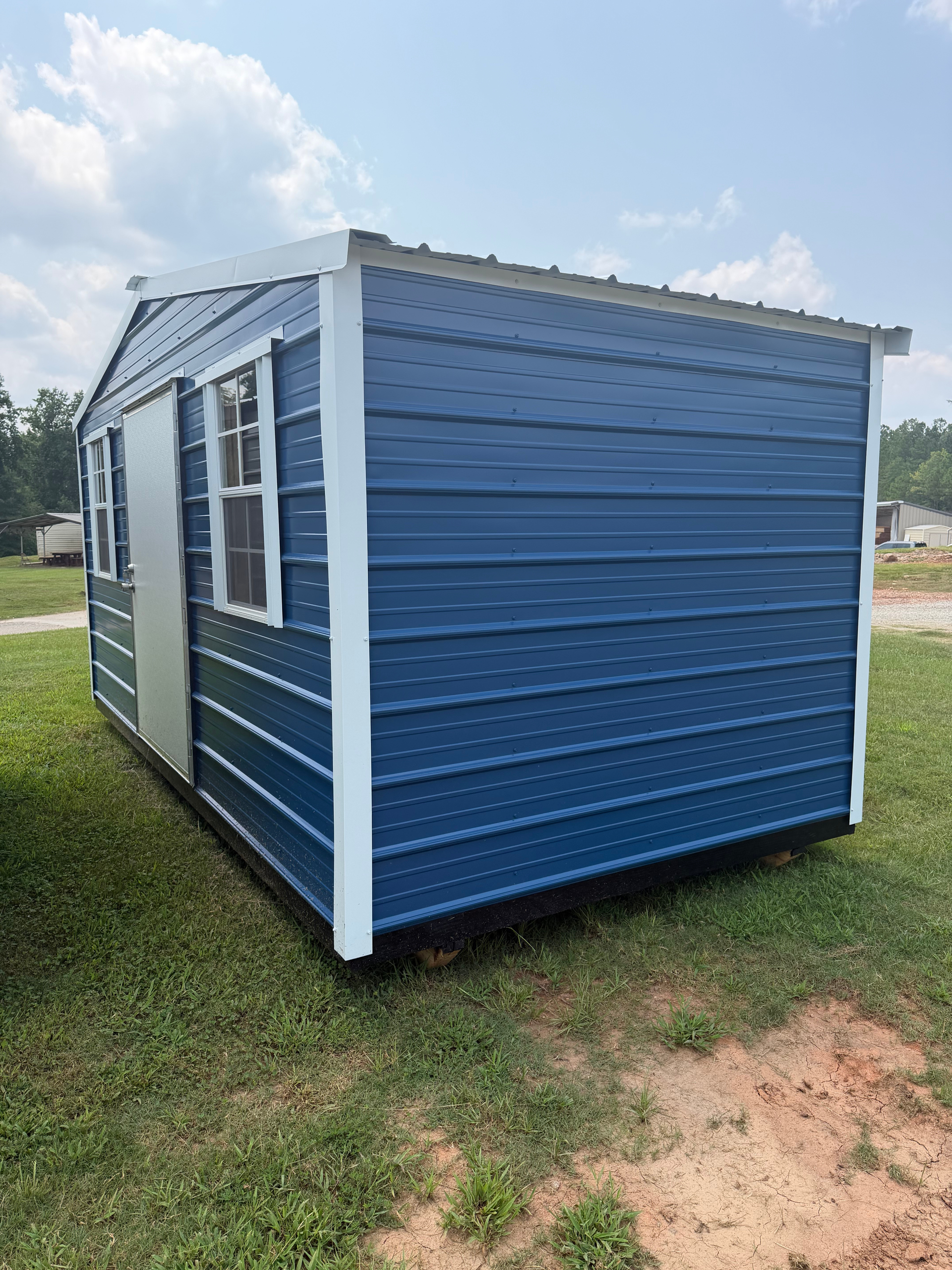Blue portable shed with white trim on a grassy lot under a partly cloudy sky
