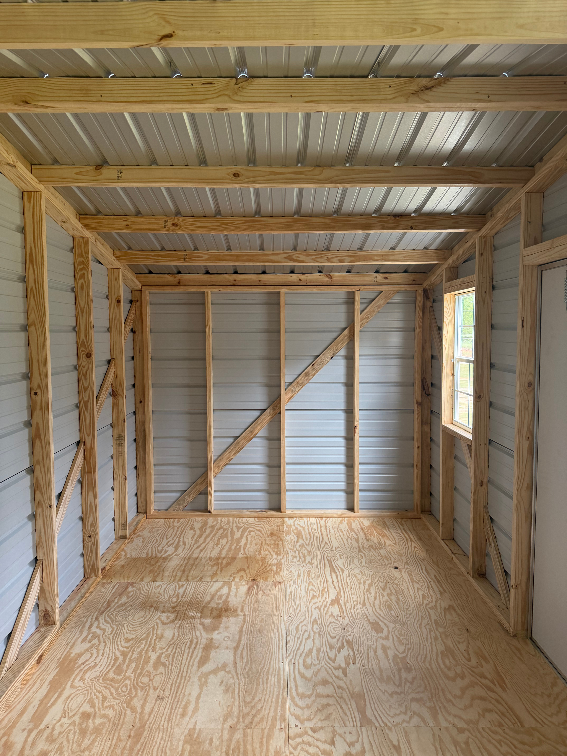 Empty unfinished wooden shed interior with plywood floor, exposed framing, and small side window.
