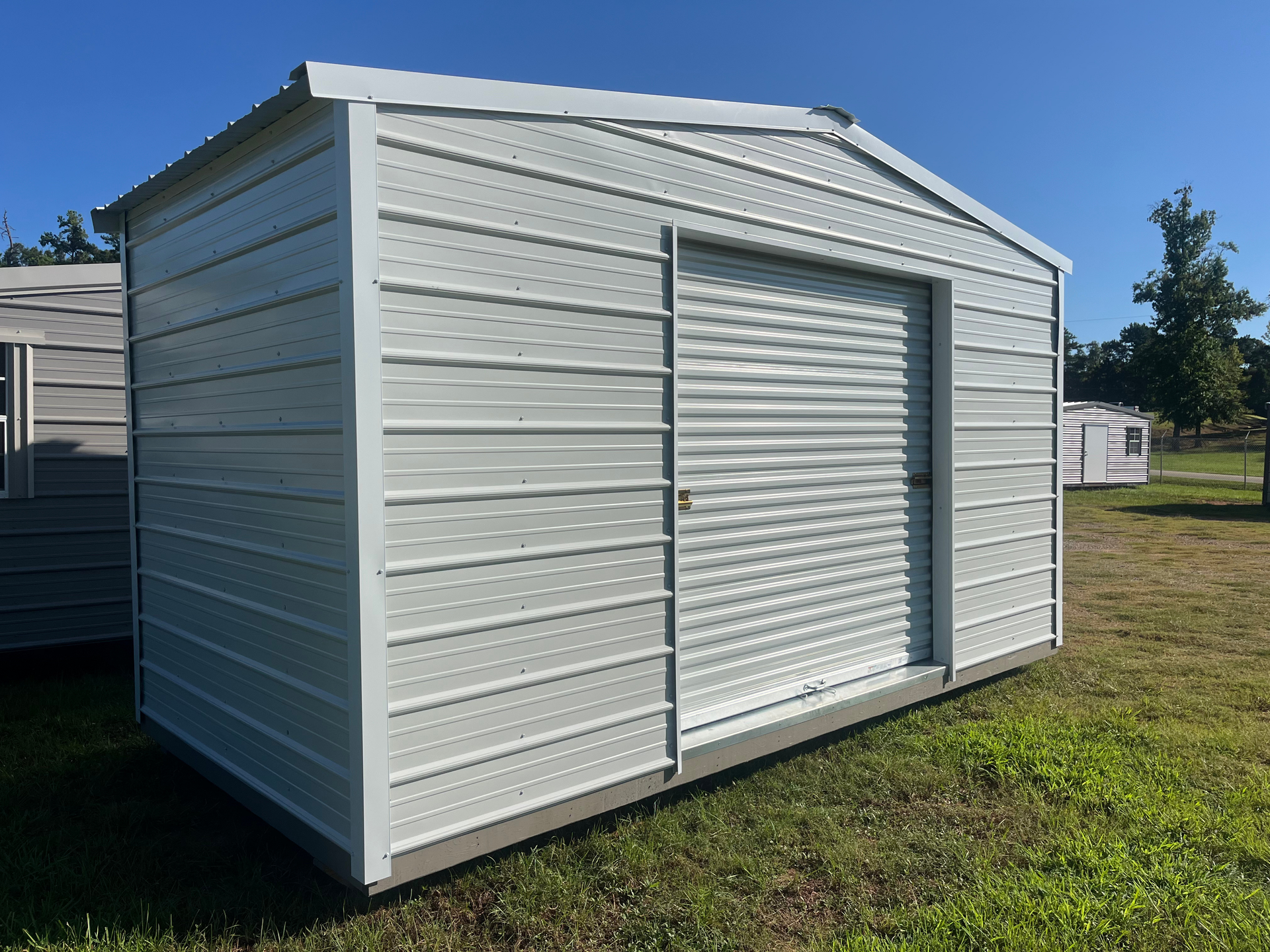 White metal storage shed with a roll-up door on grass under a blue sky