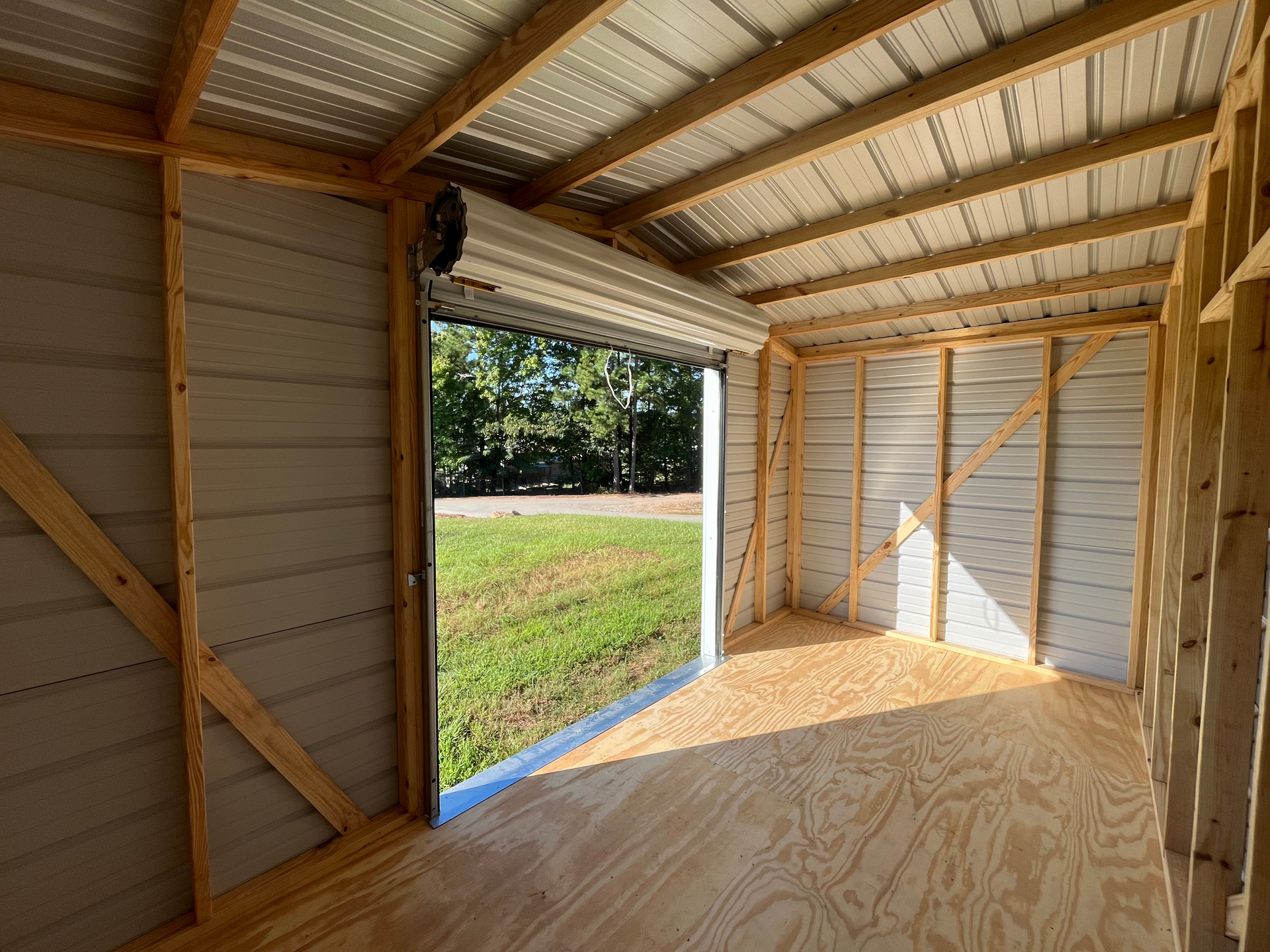 Interior of a small wooden shed with open roll-up door, plywood floor, and grassy yard outside
