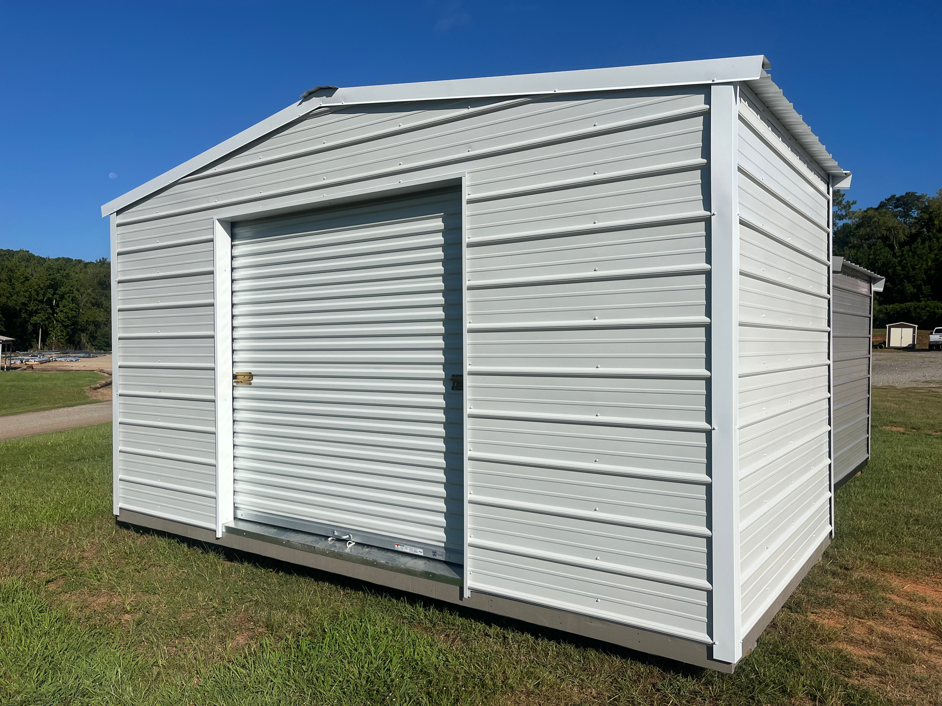 White metal storage shed with a roll-up door on a grassy lot under a clear sky