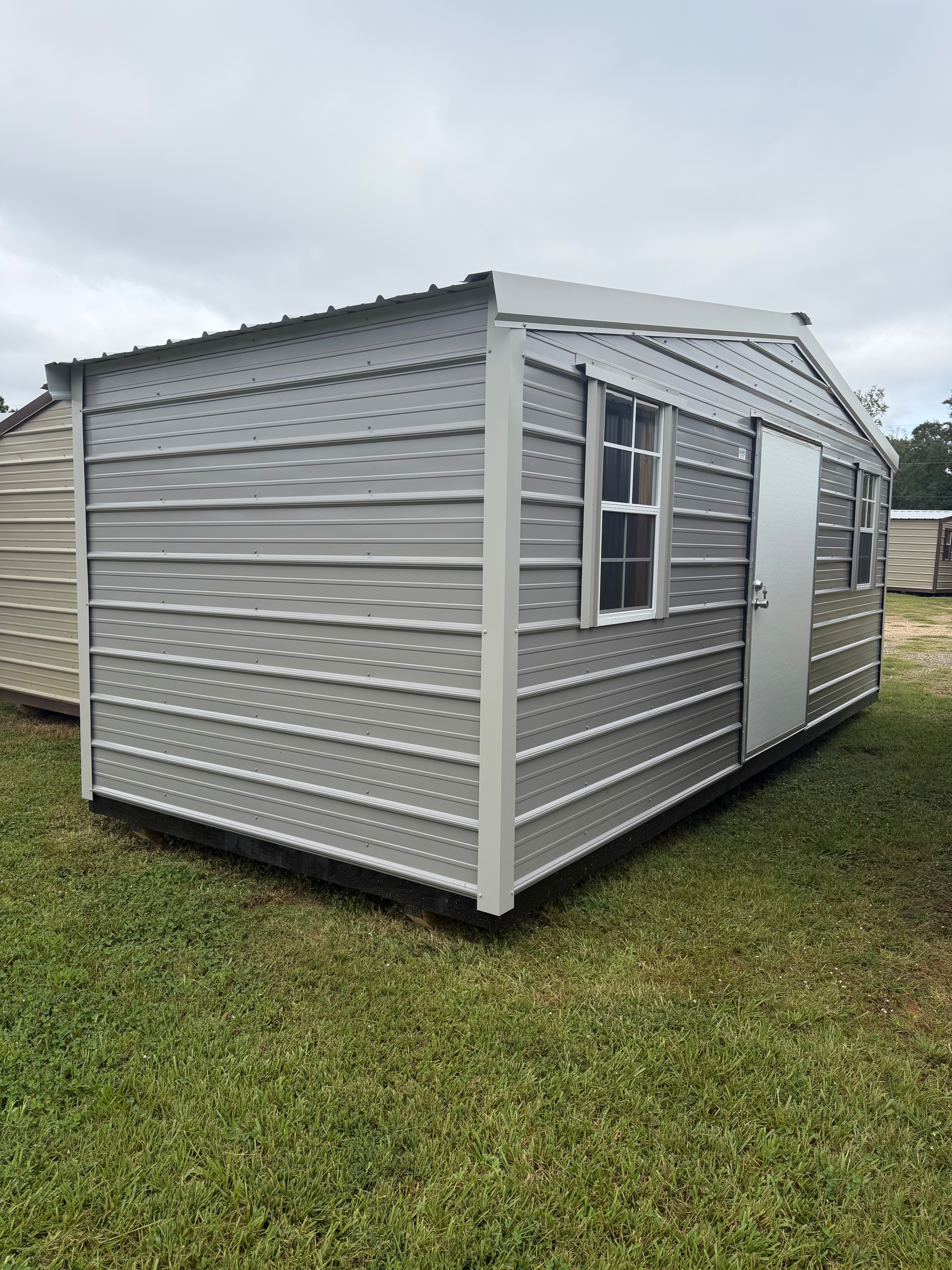 Gray portable cabin with windows and a white door on a grassy field under cloudy skies