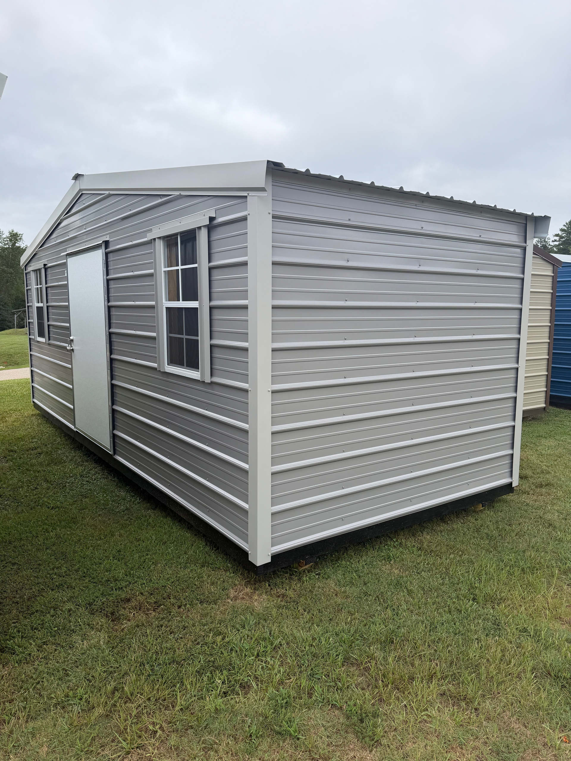 Small gray metal shed with a sloped roof and door, sitting on grass under a cloudy sky.