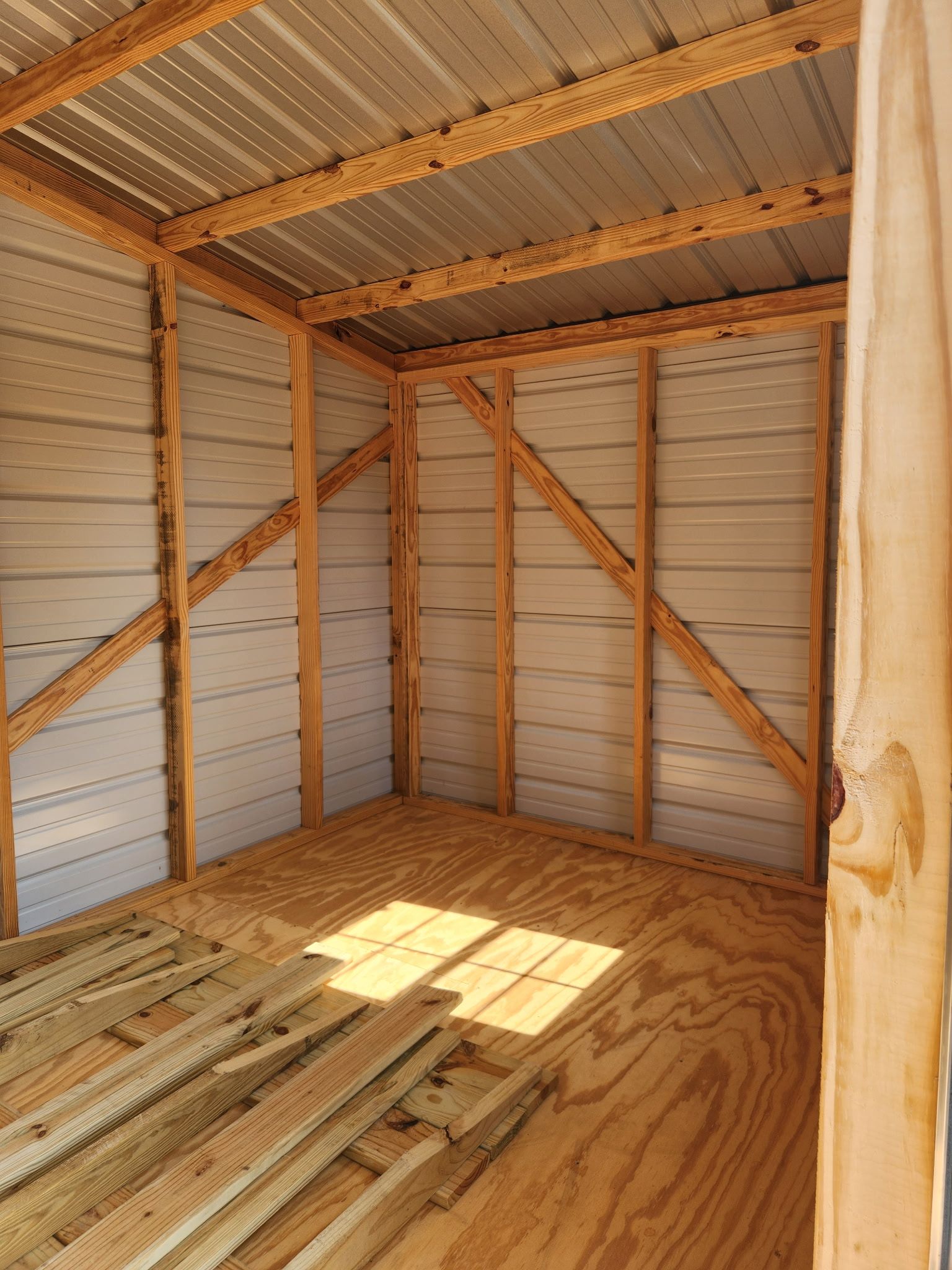 Empty wooden shed interior with exposed framing, corrugated metal roof, and stacked lumber on the floor.