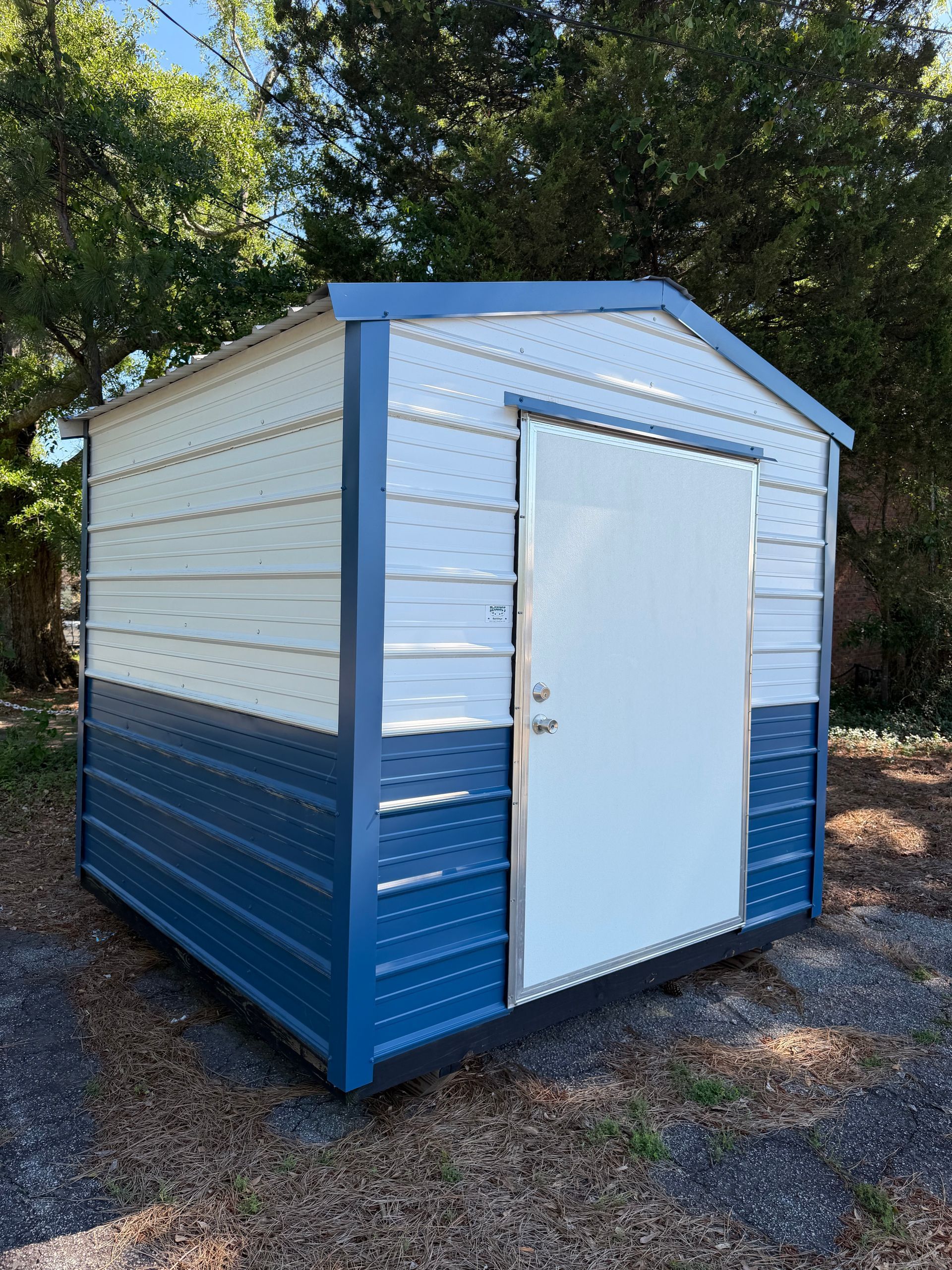 Blue-and-white metal storage shed with a white door on a gravel lot under trees