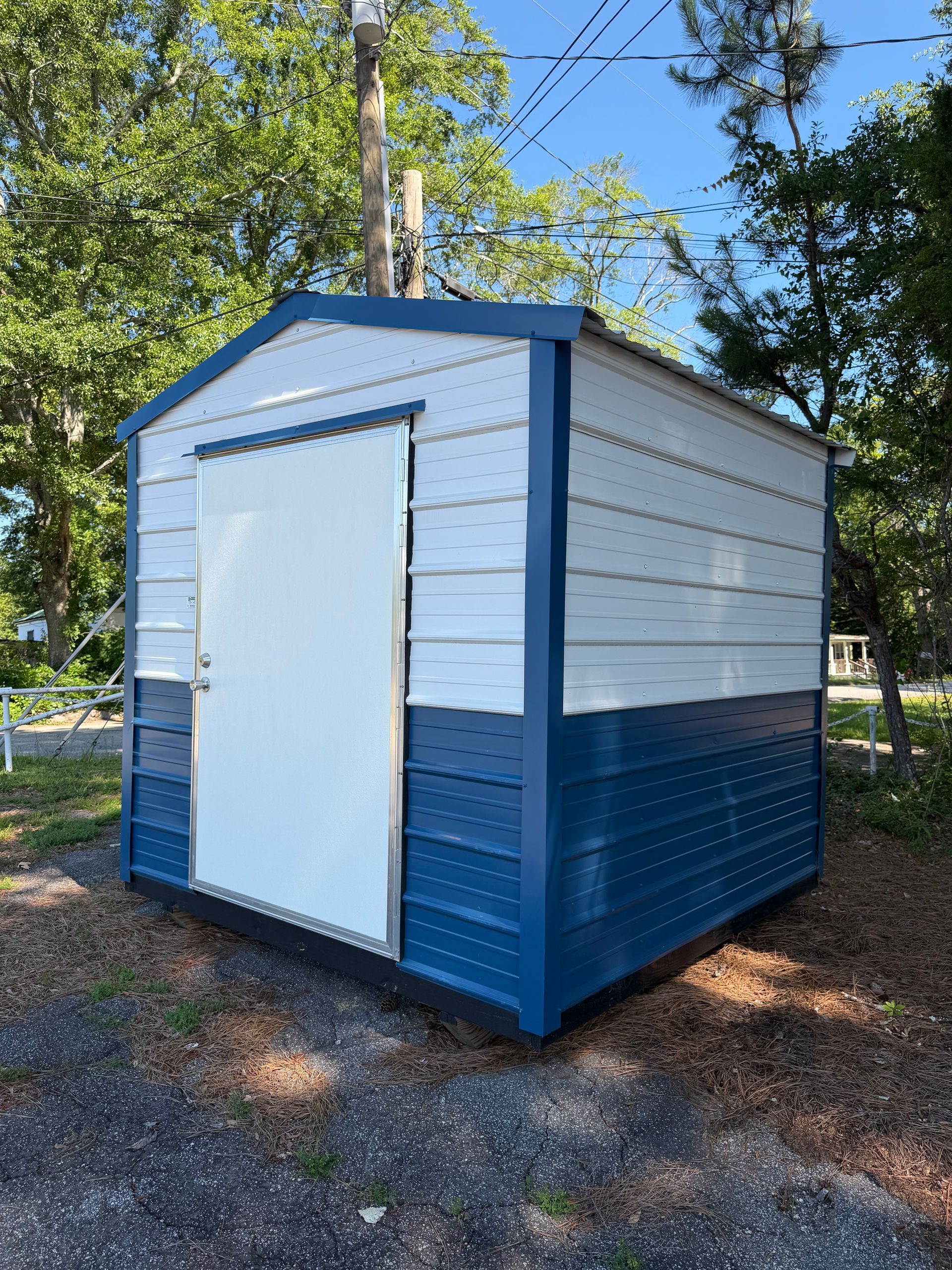 Blue-and-white metal storage shed with a white door on a gravel lot under trees