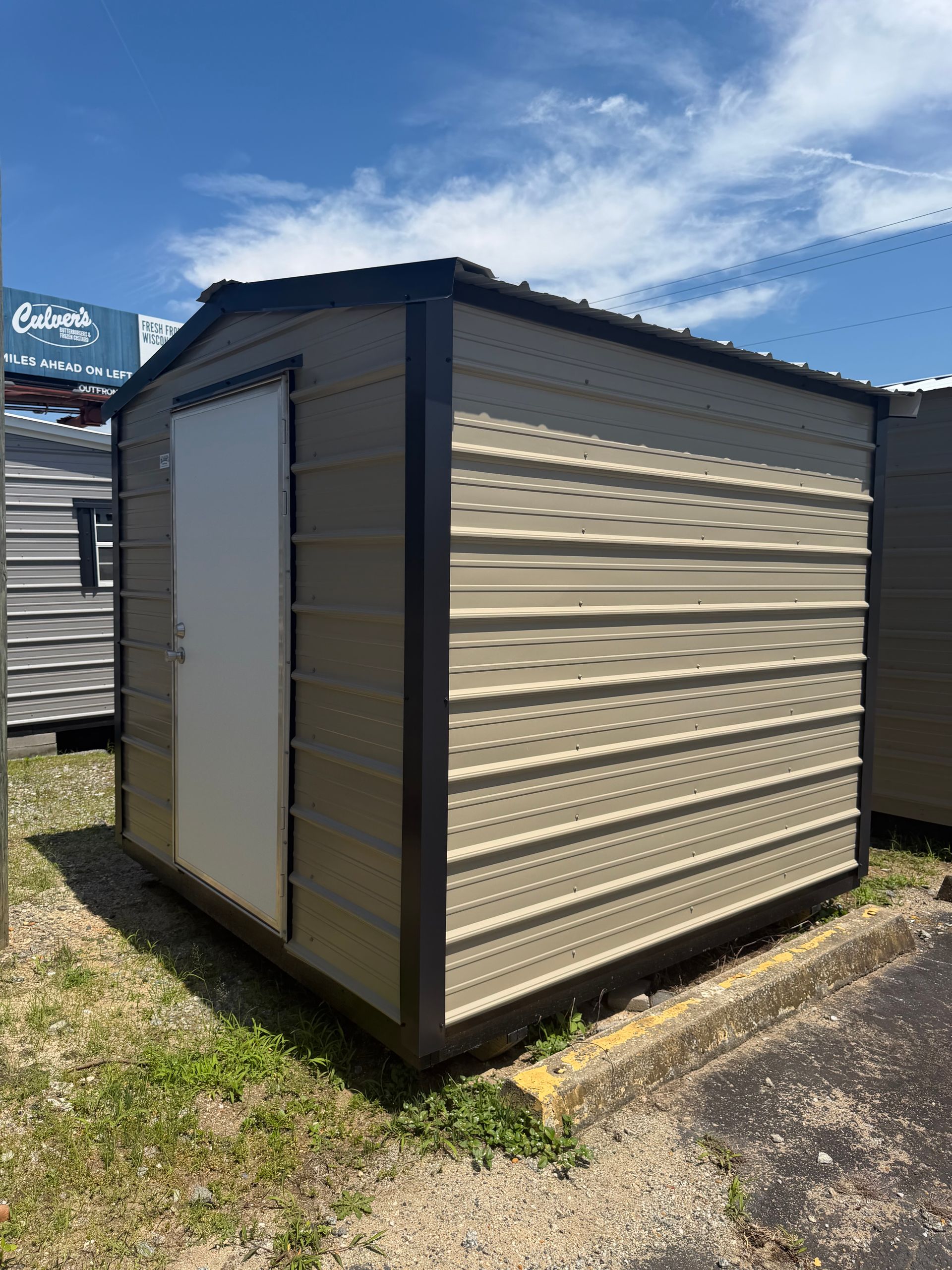 Small beige metal storage shed with a white door, sitting on grass under a blue sky.