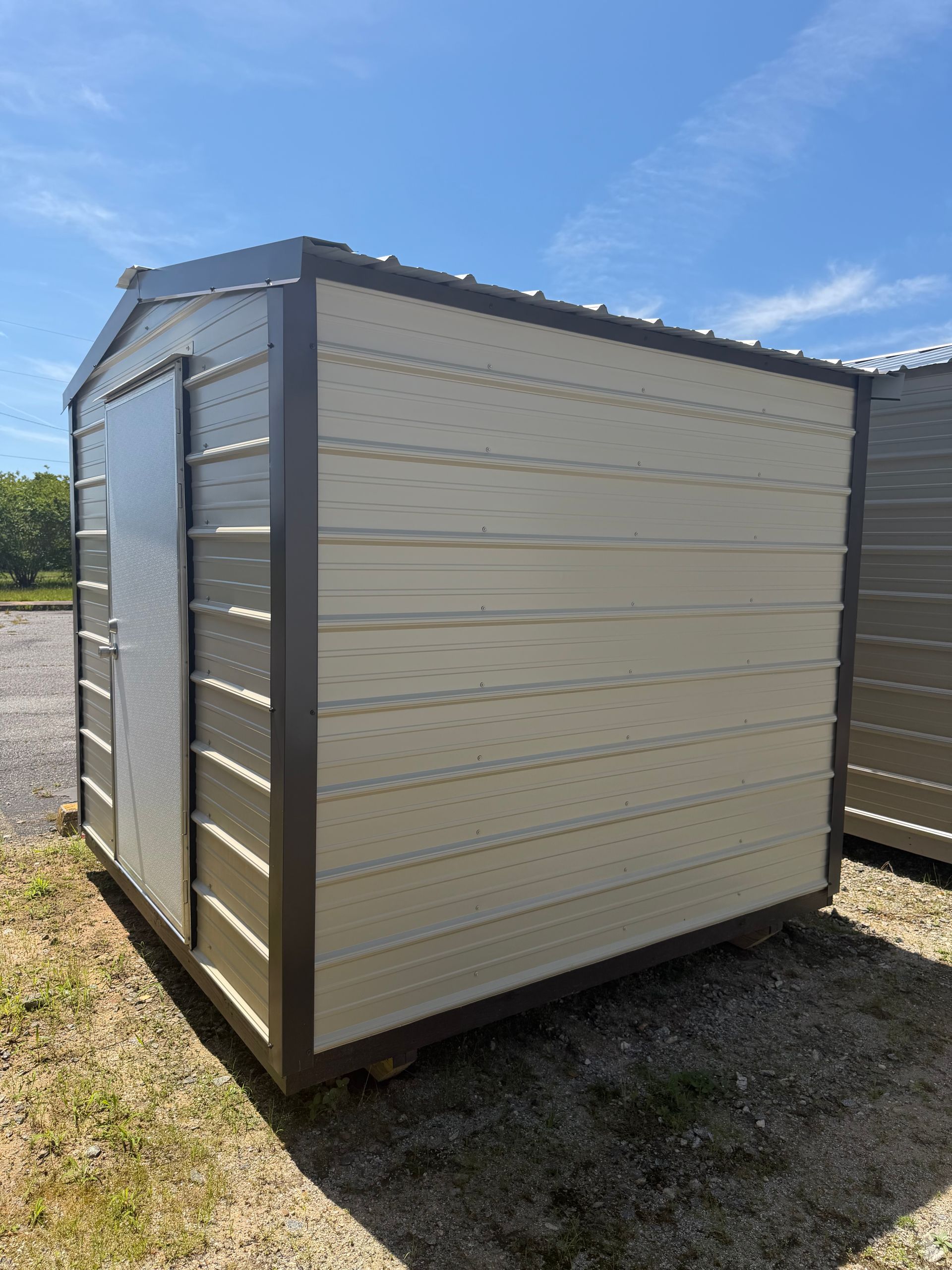 Small tan storage shed with a gray roof sitting on a grassy lot under a blue sky